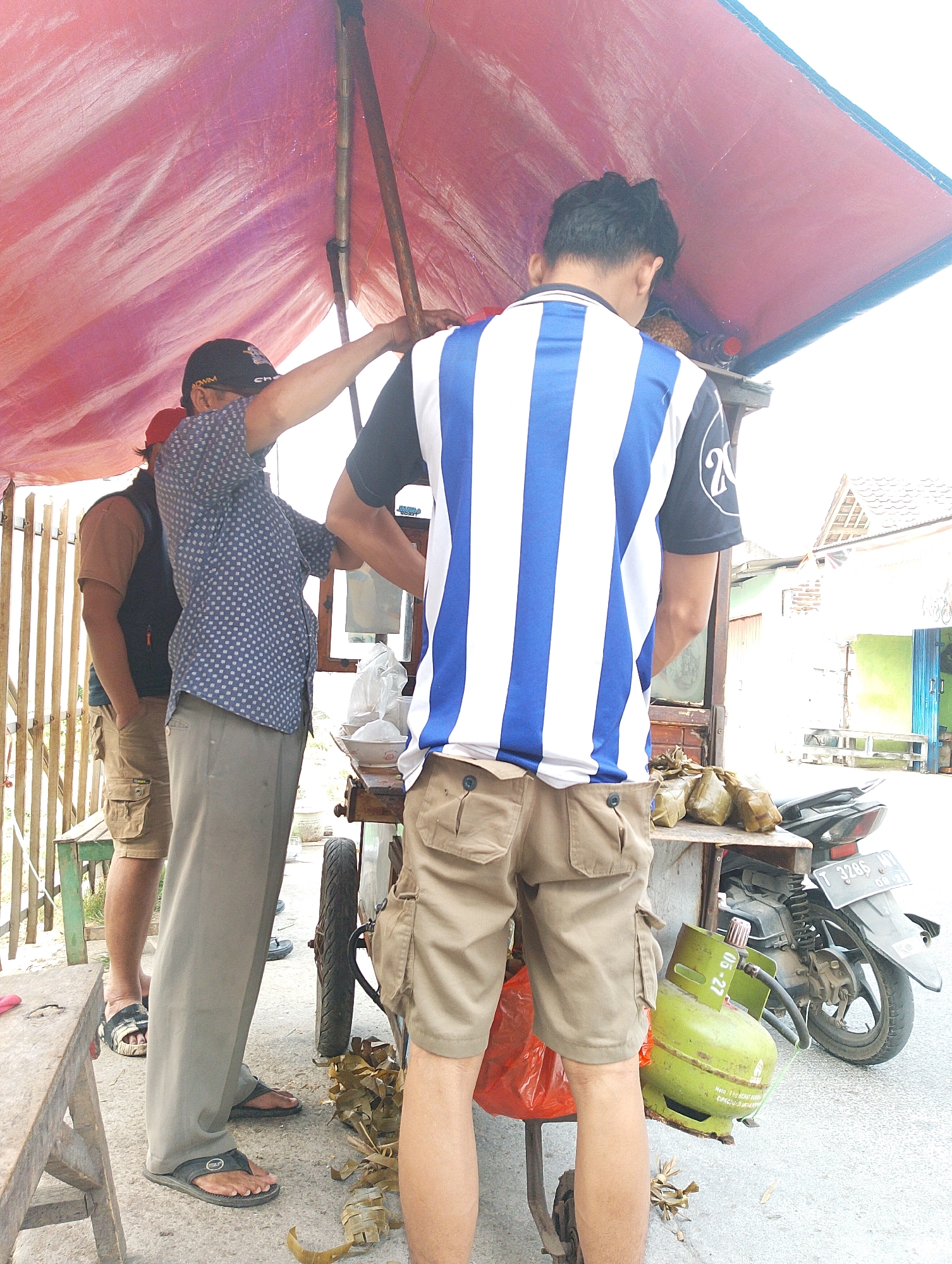 In the city of Karawang, Indonesia, under a pink tarp, a group of individuals stand around a food stall. One man, wearing a blue and white striped shirt and tan cargo shorts, stands facing away from the camera, with his back to a food cart. A second man, wearing a blue patterned shirt and grey pants, is also partially visible, his arm extended upwards. A third man, wearing a brown shirt and tan cargo shorts, stands to the left, his hand in his pocket. In front of the food cart, a green propane tank is visible. A motorcycle is parked behind the food cart. The scene appears to be outdoors, on a paved surface, with a wooden fence in the background.