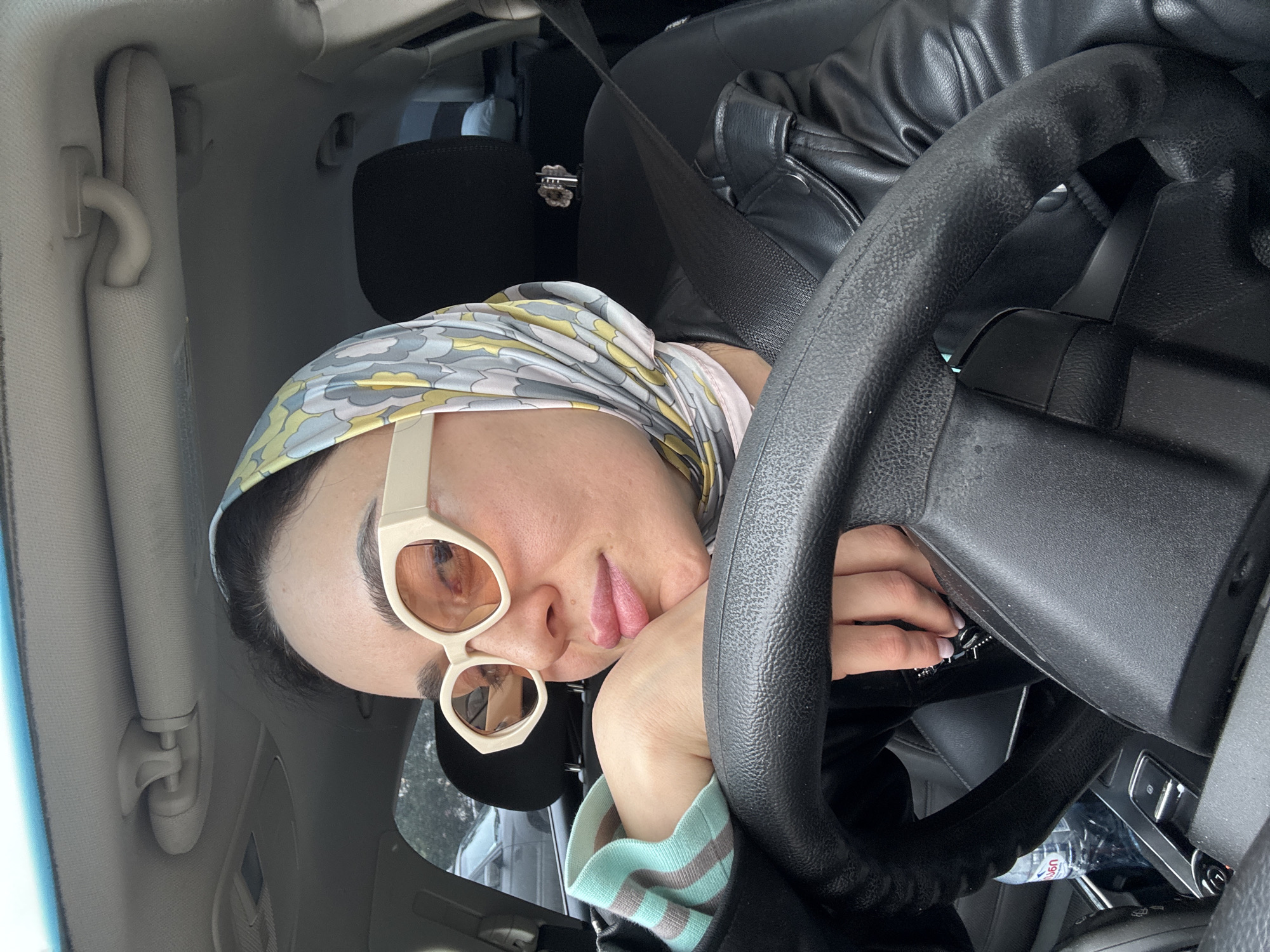 A woman is depicted resting in the driver's seat of a car, captured from a slightly high, tilted perspective. Her head is tilted and rests on her right hand, which is draped over the black, textured steering wheel. She wears a patterned headscarf featuring abstract shapes in yellow, light blue, grey, and pink. Large, off-white, cat-eye style sunglasses with amber-tinted lenses cover her eyes, reflecting indistinct shapes of the environment. Her lips are slightly parted, revealing a hint of her tongue, conveying a relaxed or tired expression. She is dressed in a dark jacket, possibly leather or faux leather, and a light-colored top with teal/mint green and grey striped cuffs. Her left hand, with manicured nails, is visible near the steering column.
The car's interior is visible, including a black seatbelt crossing her chest, part of a black car seat, and the light grey/beige headliner with a sun visor and grab handle. Below the steering wheel, a plastic water bottle with a blue cap is partially visible, with the numbers "690" clearly legible on its label. The lighting is soft and diffused, suggesting it is daytime. Her posture and expression convey a sense of calm, rest, or contemplation. The "690" on the water bottle is a common barcode prefix for products from Georgia, providing a specific location cue consistent with Tbilisi, Georgia.
