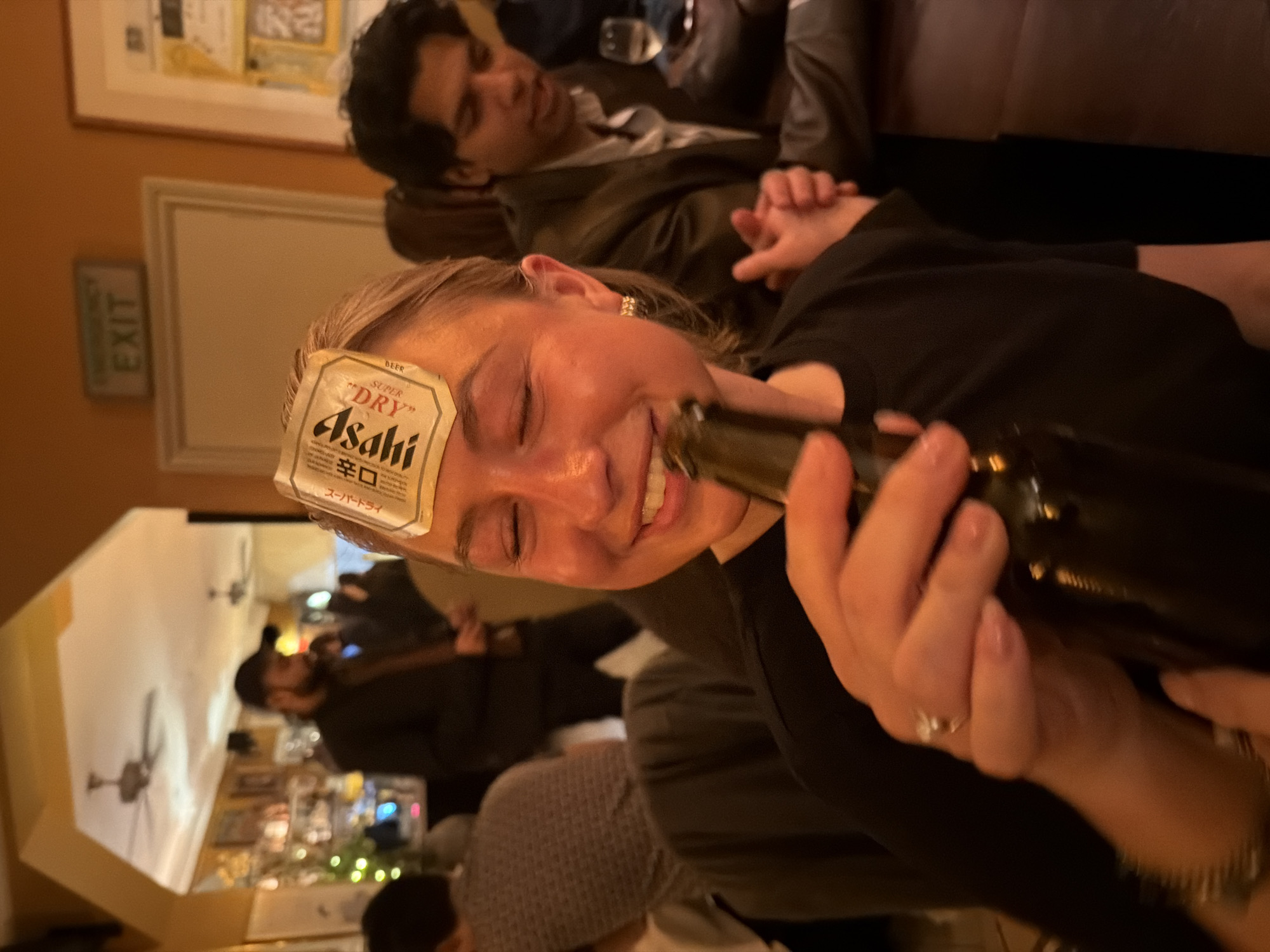 In a dimly lit interior scene, a smiling woman with a beer label on her forehead holds a dark bottle in her hands. She is wearing a dark top and a ring. Behind her, another person is lying down, dressed in a jacket and a light-colored shirt. Other individuals and a bar area with drinks and decorations are visible in the blurred background. The presence of an "EXIT" sign suggests a public establishment. The location appears to be London, United Kingdom.