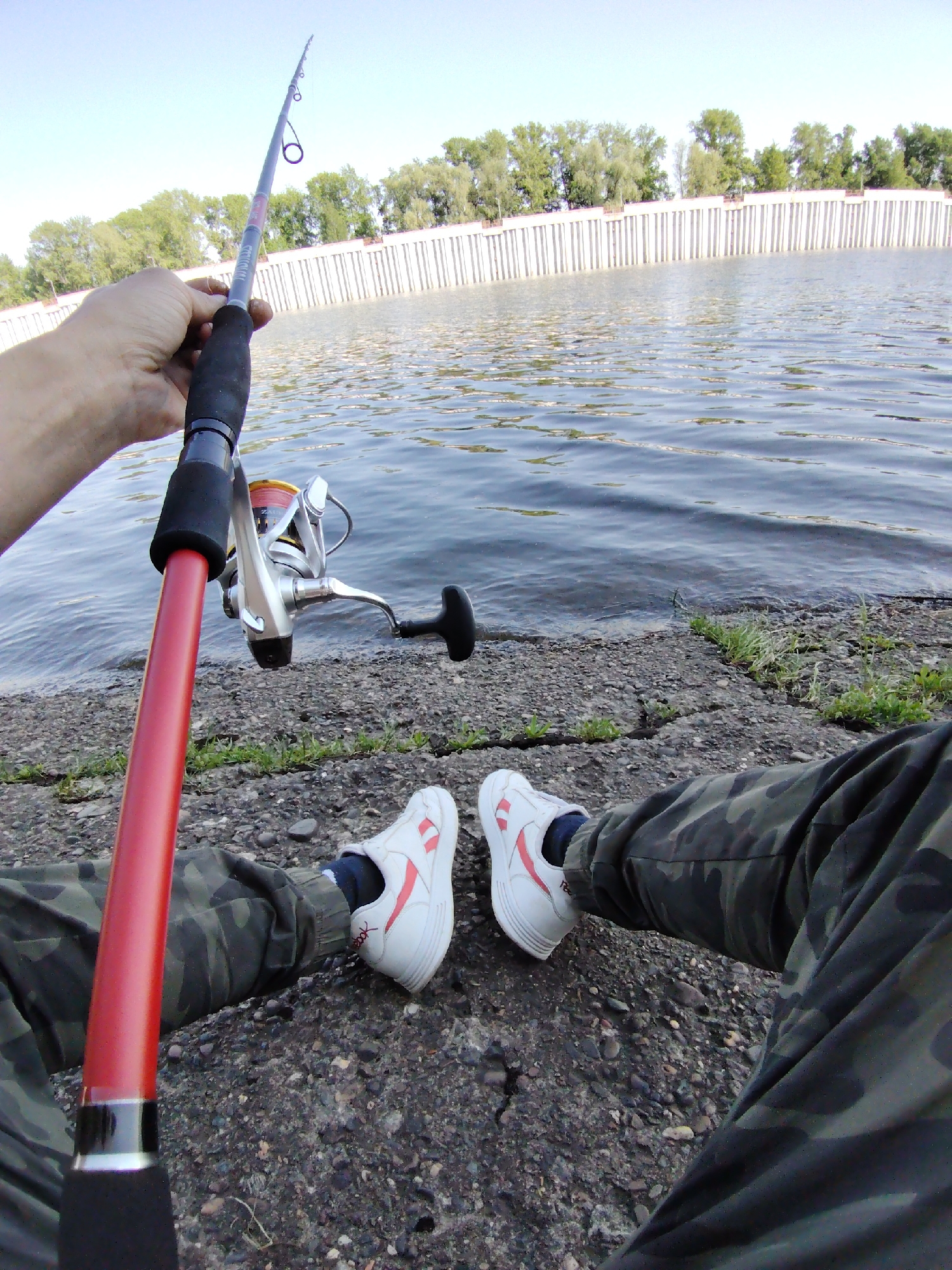 An individual sits on a rocky embankment by a body of water, holding a red fishing rod. The water is calm with gentle ripples. Beyond the water, a long, white concrete structure lines the horizon, topped with a row of green trees. The individual is wearing camouflage pants and white sneakers with red accents. The location is in the city of Chaikovsky, Russia.