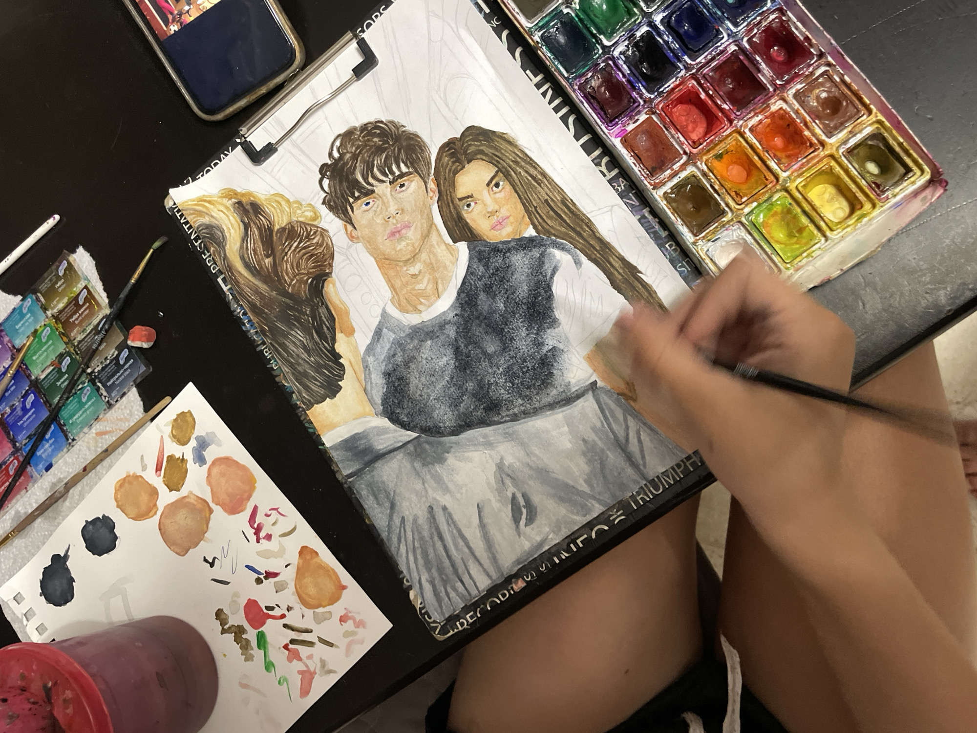 An overhead, medium shot captures a person's hands and arms as they paint a watercolor portrait of two individuals. The artwork, a detailed rendering of a man and a woman, is laid out on a black surface, possibly a table or desk. The woman's hair is long and brown, cascading down her shoulder. The man's hair is also brown and appears to be styled in loose curls. They are both looking towards the viewer with soft expressions.
Beside the painting, a tray of watercolor paints is visible, with various hues of red, orange, yellow, green, blue, and brown. Brushes and a small white eraser are also present. To the left of the painting, a white sheet of paper displays splotches of various colors, likely test swatches from the watercolor set. A pinkish-red container, possibly for water or mixed paint, stands to the left of this swatch paper. A smartphone with a dark blue case is also on the black surface to the right of the painting. The overall environment suggests an art studio or a creative workspace. There is no indication of the specific location within the city of Sochi, Russia.