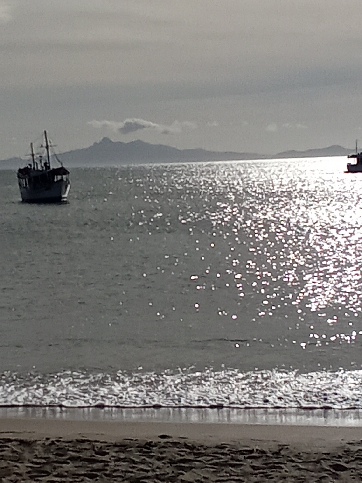 The image depicts a coastal scene in Juan Griego, Venezuela. A body of water, likely the Caribbean Sea, dominates the view, with sunlight reflecting off its surface. A fishing boat is anchored to the left of the frame. A second boat is located further to the right and in the background. Mountains are visible on the horizon behind the boats. The foreground shows a sandy beach with waves. The sky appears overcast.