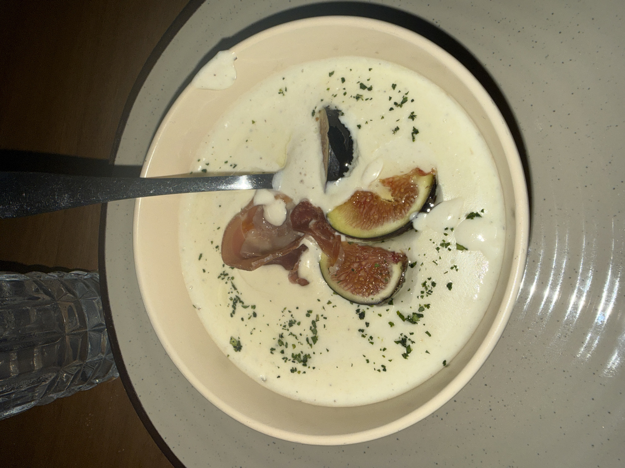 The image shows a bowl of soup, topped with figs and ham, on a plate. A spoon is in the bowl. A textured glass is visible in the lower-left. The scene is lit with artificial light. The location is Unknown, Unknown.