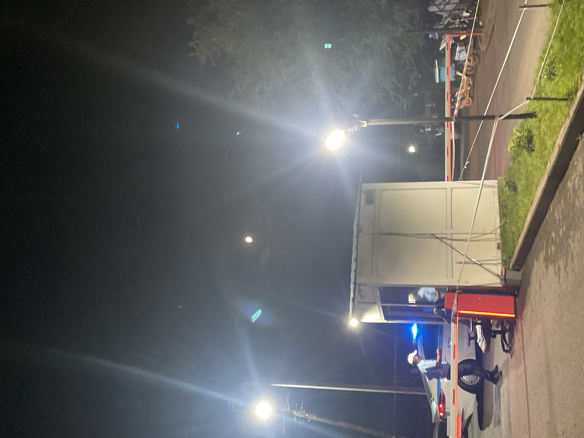 The image depicts a nighttime scene at what appears to be a security checkpoint, possibly at the entrance to an industrial area or a gated community in Almaty, Kazakhstan. A white security booth is positioned near a lowered barrier gate, which is illuminated with red and white stripes. Two individuals in uniform, wearing white caps, are present. One individual is standing next to a white vehicle with its emergency lights flashing blue. This individual appears to be interacting with someone inside the security booth. The second uniformed individual is visible inside the security booth, appearing to be on duty. The environment is dimly lit, with overhead streetlights casting bright beams and creating lens flares. Trees are visible in the background, and grass and pavement make up the immediate surroundings. Several motorcycles are parked beyond the barrier gate. The sky is dark with a faint moon visible.
