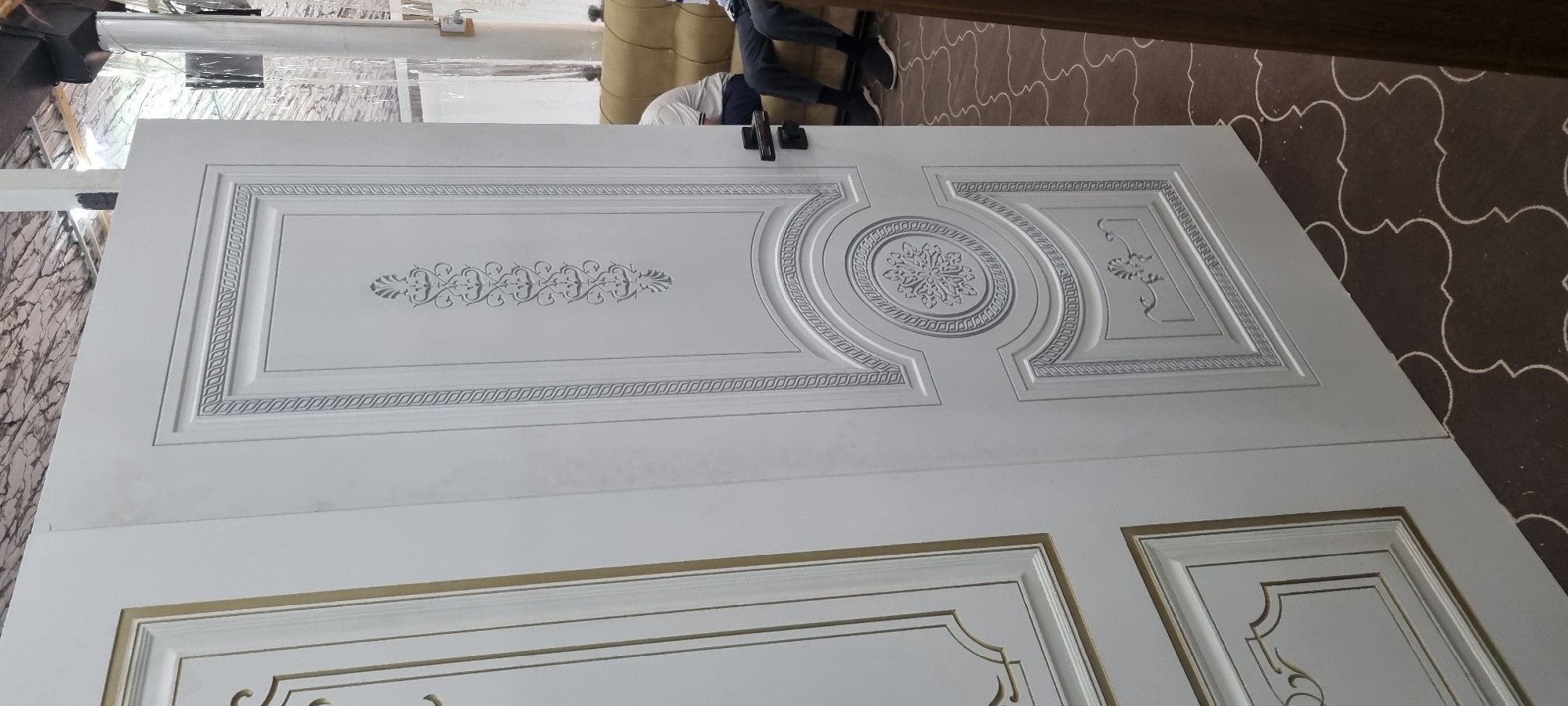 In Bokhtar, Tajikistan, an interior scene features two white doors with intricate decorative molding. The door on the right is the primary focus, displaying a central rectangular panel with a symmetrical, carved floral design. This panel is surrounded by a rope-like textured border and further enclosed within an arched frame, also decorated with the rope texture. Above this, a vertical panel showcases a repeated, stylized leaf or feather motif. Below the central arched panel, another rectangular panel features a simpler, curved floral element. The door on the left, partially visible, has a similar style, with decorative panels outlined in gold trim. The floor is covered in a dark brown carpet with a repeating white clover-like pattern. In the background, a tan sofa is visible, with two individuals seated on it, partially obscured. One person wears a white shirt and dark pants, while the other wears a light-colored shirt and dark pants. They appear to be passively present in the scene. The lighting suggests an indoor environment with overhead illumination.
