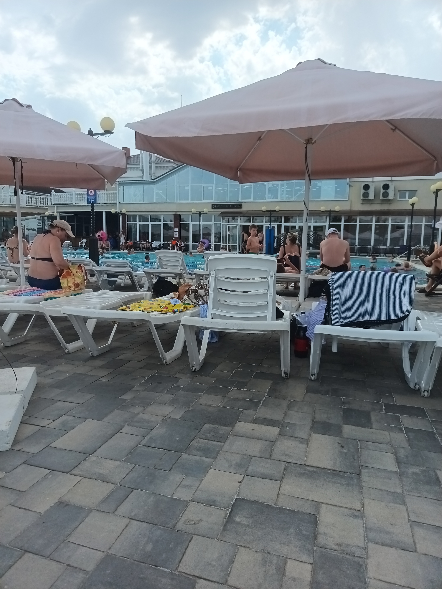 The image shows a poolside scene with several lounge chairs in the foreground and a swimming pool in the background. People are scattered around the pool area, some swimming, others relaxing on lounge chairs or standing. Large beige umbrellas provide shade over some of the chairs. The ground is paved with rectangular stones. A building with large windows is visible behind the pool. The sky is cloudy.