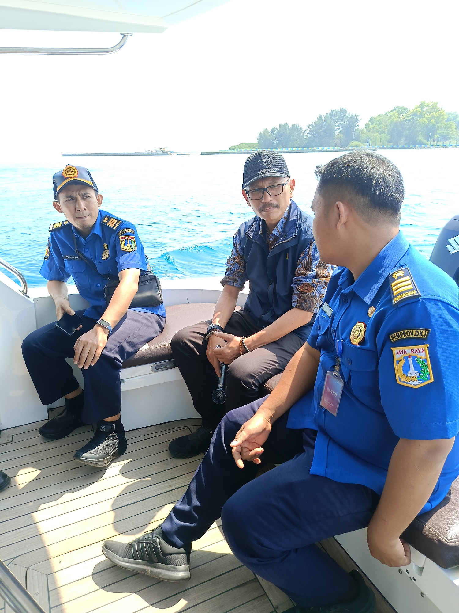 Three men in blue uniforms are seated on a boat on the water. The man on the left is wearing a blue uniform shirt with gold insignia on the shoulders and a blue cap with a gold emblem. He is holding a smartphone in his left hand and has a black crossbody bag slung over his shoulder. The man in the middle is wearing a dark blue vest over a patterned shirt and a black baseball cap. He is wearing glasses and has a mustache. The man on the right is wearing a blue uniform shirt with gold insignia on the shoulders and a patch on the left breast with "PEMPROV.DKI JAYA RAYA" and an emblem of Jakarta. He is looking towards the man in the middle. The boat has a wooden deck. In the background, there is blue water and a distant island with trees. The scene appears to be in the waters surrounding Jakarta, likely in the Thousand Islands (Kepulauan Seribu) area, a popular tourist destination and part of the Special Capital Region of Jakarta, Indonesia.