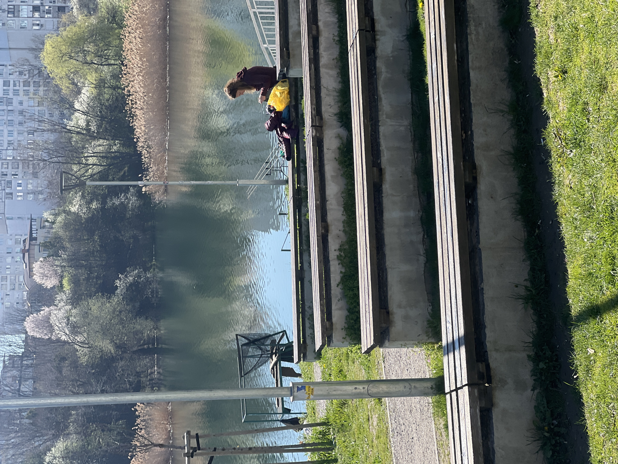 The image depicts a waterside scene in Sofia, Bulgaria, with a canal-like body of water flanked by concrete and wooden stepped platforms on one side and a grassy bank on the other. A woman is sitting on the steps with a yellow bag. The water reflects the surrounding buildings, trees, and reeds. Metal poles and a metal structure are visible near the water's edge. The scene is bathed in daylight.