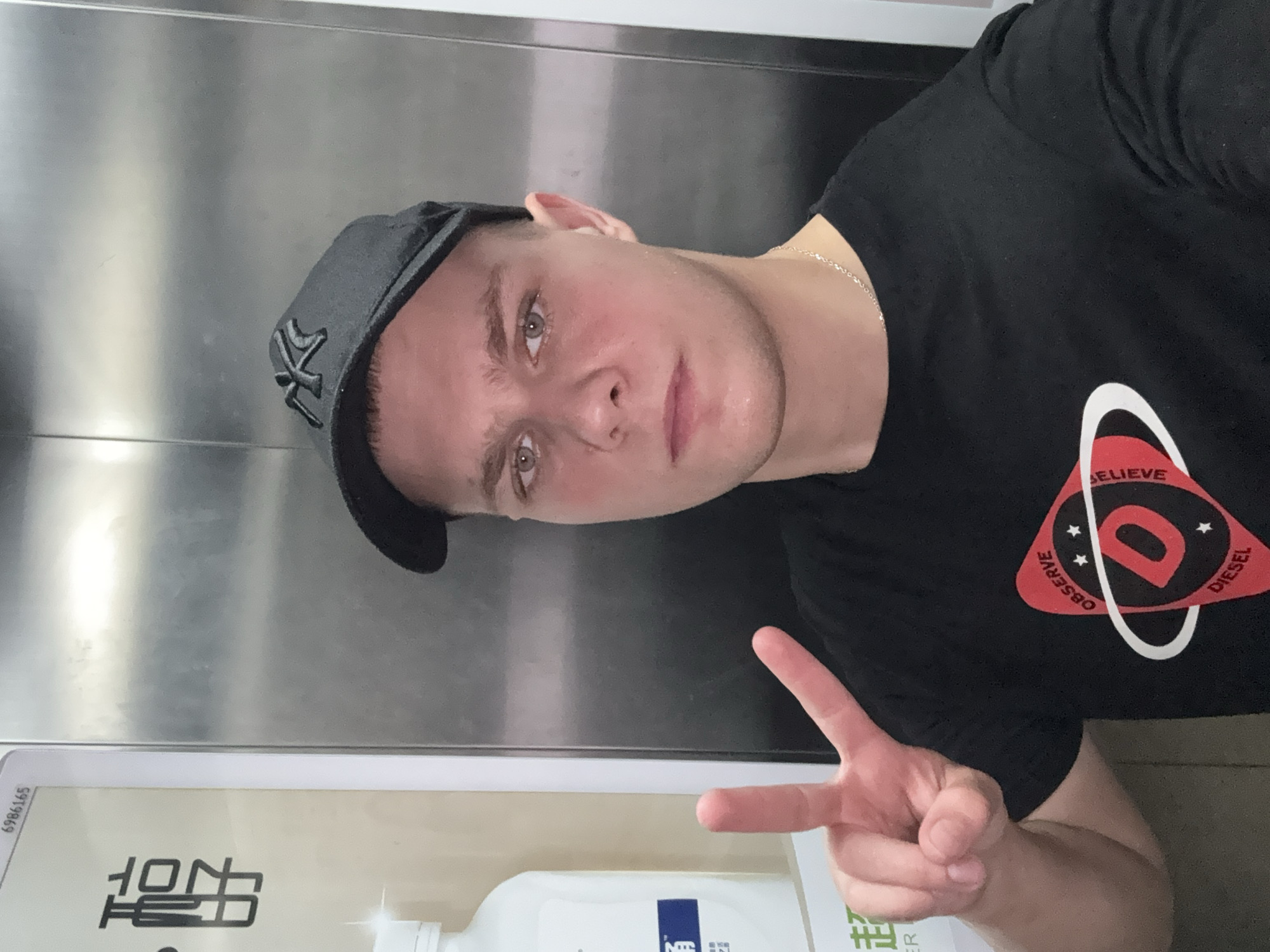 In Haizhu District, China, a young Caucasian male with light skin, light brown hair, and blue eyes is pictured in an elevator. He is wearing a black baseball cap with a grey logo, a black t-shirt with a circular red and white graphic that says "OBSERVE, BELIEVE, DIESEL," and a silver chain necklace. He is looking at the camera, holding up his right hand with his index and middle fingers extended in a peace sign. To his left, a white bottle with a blue label is visible. The background is a brushed metal elevator wall. The image appears to be a selfie taken in an elevator.