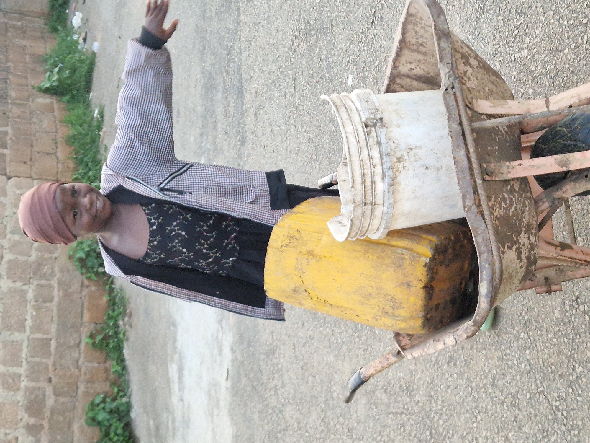 An outdoor medium shot captures a young Black girl standing beside a rusty wheelbarrow filled with two buckets, one yellow and one white. The girl wears a brown headscarf, a patterned dress, and a checkered jacket, and she is extending her right arm outward. The wheelbarrow is positioned on a gravelly surface, with a brick wall and sparse vegetation in the background. The lighting suggests it is daytime.