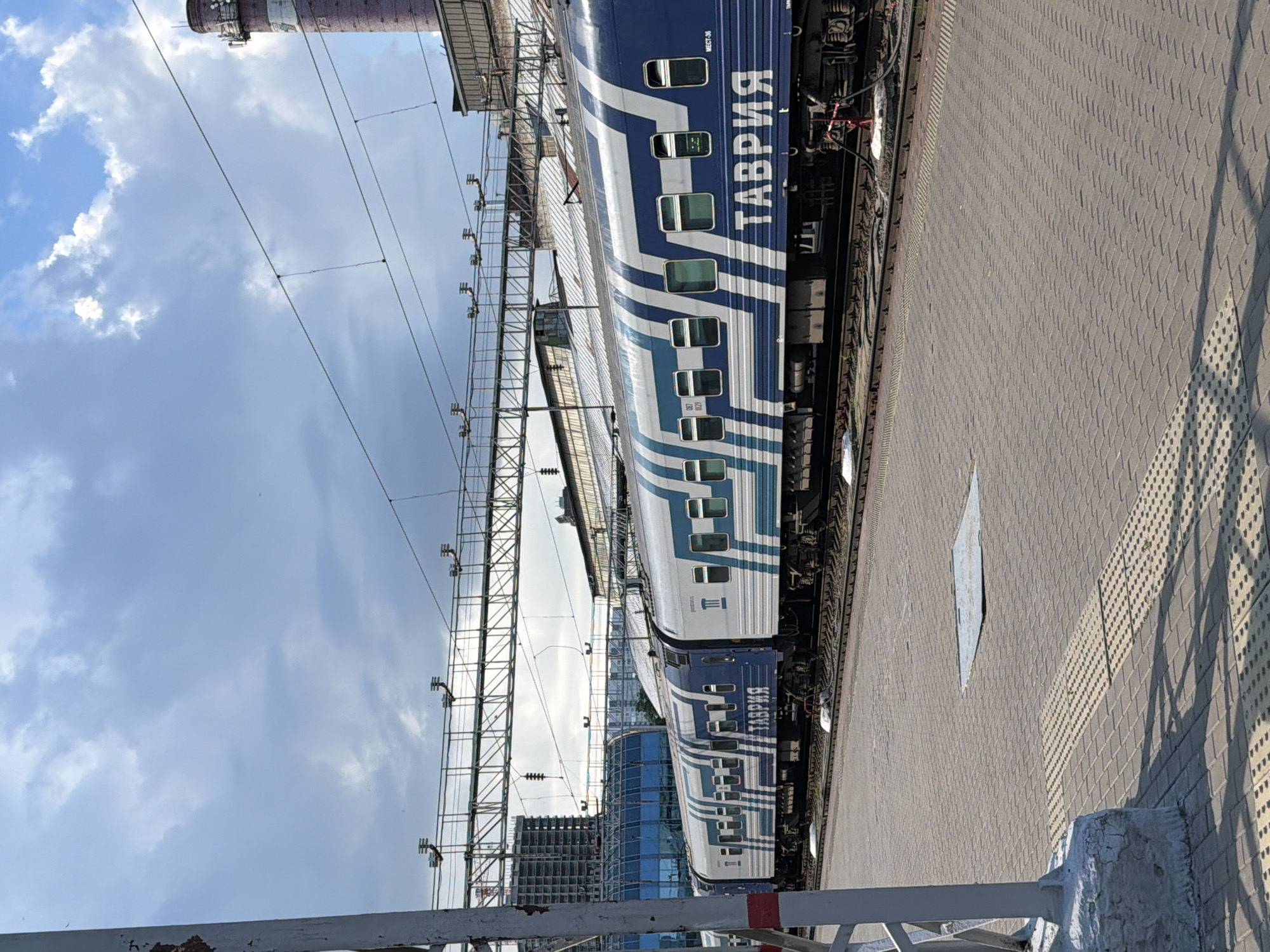 The image shows a train at a station in Rostov-on-Don, Russia. The train cars are blue and white with a distinctive striped pattern. The word "ТАВРИЯ" (TAVRIYA) is written in large white letters on the side of one of the cars. The train is on a track, with a paved platform to the right. In the background, there are overhead electrical wires, a tall brick chimney with antennas, and a partially constructed building. The sky is partly cloudy. The overall scene suggests a typical operational train station environment in an urban setting.