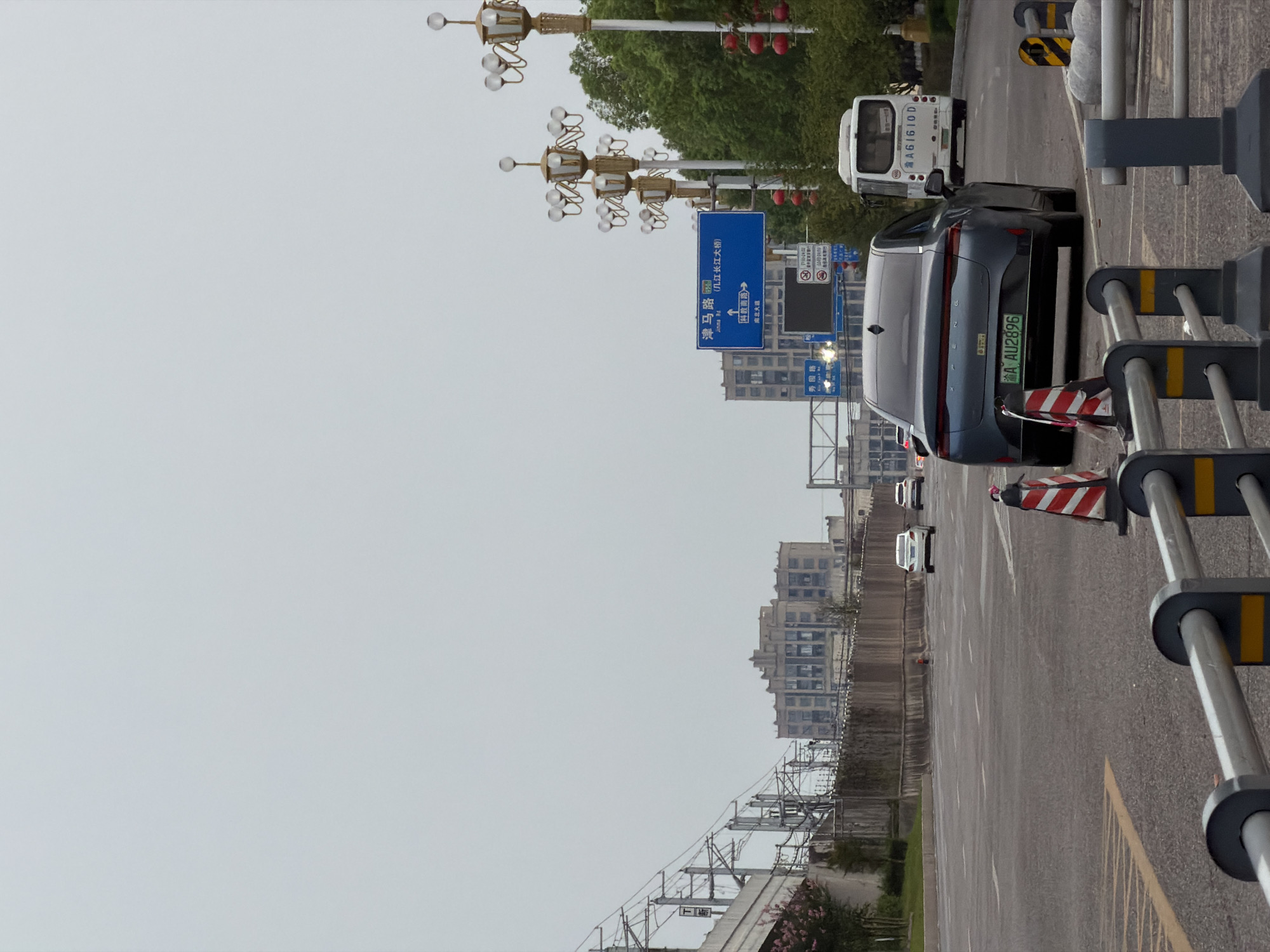 A grey sedan with license plate AU2896 is parked on an asphalt road in Jiangjin, China. The car is facing away from the viewer, and traffic cones are placed in front of it. A white bus with license plate AG1610D is parked behind the sedan. The sky is overcast. In the background, there are buildings, trees, and streetlights. A railway line is visible on the left side of the image. A blue directional sign indicates "Jingma Road" and "Yingde Bridge".