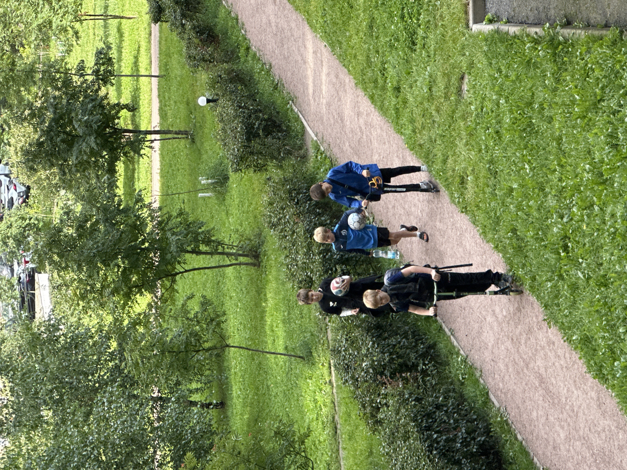 Four boys are walking and riding scooters on a gravel path through a park in Murino, Russia. The boy on the left is on a scooter and wearing a dark blue vest over a black t-shirt and black pants. The boy next to him is wearing a black t-shirt and black shorts and holding a soccer ball. The third boy from the left is wearing a blue shirt and black shorts and holding a soccer ball and a plastic water bottle. The fourth boy is on a scooter and wearing a blue jacket and black pants. The path is lined with green bushes and grass. Trees are visible in the background, along with parked cars and buildings. The scene appears to be from a residential area.