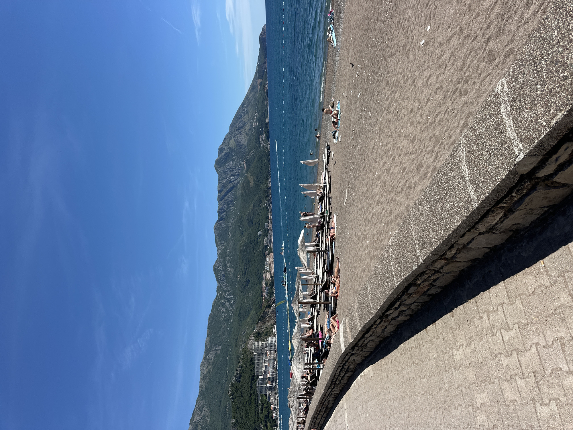 This is a medium-angle, full-shot, eye-level photograph taken on a clear, sunny day in Boreti, Montenegro. The scene captures a bustling beach resort area along the Adriatic Sea. The foreground features a textured paved walkway on the left, bordering a low, stone-and-concrete wall. Beyond the wall, a stretch of dark sand beach is populated with numerous lounge chairs and thatched umbrellas, many occupied by sunbathers.
In the mid-ground, the deep blue sea is visible, with a few people swimming or wading near the shore. A pier or walkway extends into the water in the distance. Further out, several boats, including a speed boat leaving a wake, traverse the water.
The background is dominated by lush green mountains that rise steeply from the coastline. Along the base of the mountains, the resort town of Boreti is visible, characterized by a cluster of tall, white buildings. The sky above is a clear, vibrant blue, with only a few wispy clouds. The overall impression is one of a vibrant and active summer day at a coastal resort.