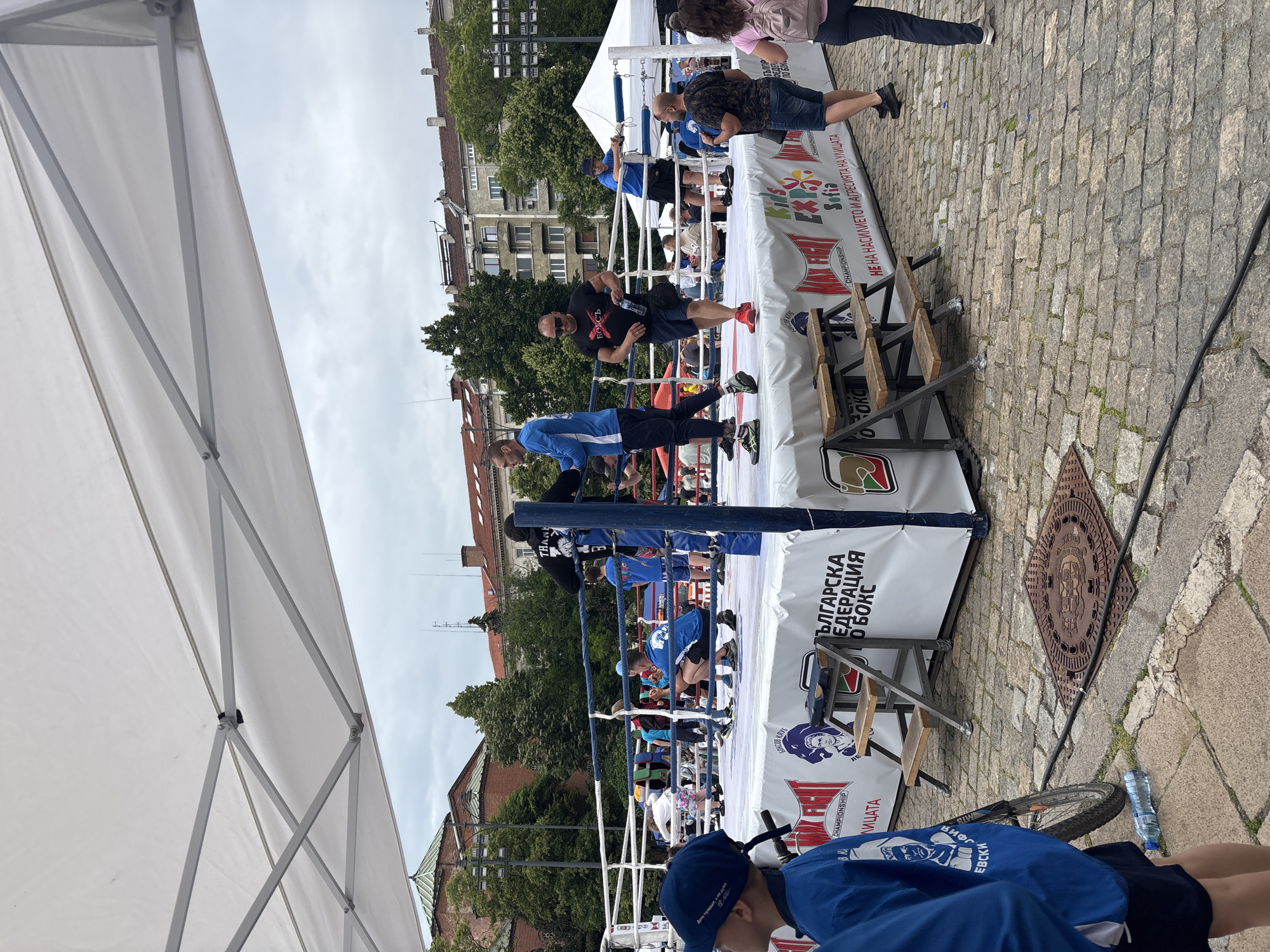 The image shows an outdoor boxing ring set up in what appears to be a public square in Sofia, Bulgaria. The ring is surrounded by spectators and coaches. A person in a black shirt with "БОКС" printed on it is inside the ring. The ring's perimeter is covered with banners in Bulgarian, with text including "БЪЛГАРСКА ФЕДЕРАЦИЯ О БОКС" (Bulgarian Boxing Federation), "CHAMPIONSHIP", "Sofia", and "НЕ НА НАСИЛИЕТО И АГРЕСИЯТА НА УЛИЦАТА" (No to violence and aggression on the street). The ground is paved with cobblestones. In the foreground, a person wearing a blue jacket is visible, with "КИФО ЕВСКИ" written on it. A bicycle and a water bottle are on the ground near the ring. Buildings and trees can be seen in the background. A white tent is partially visible on the left side of the image.