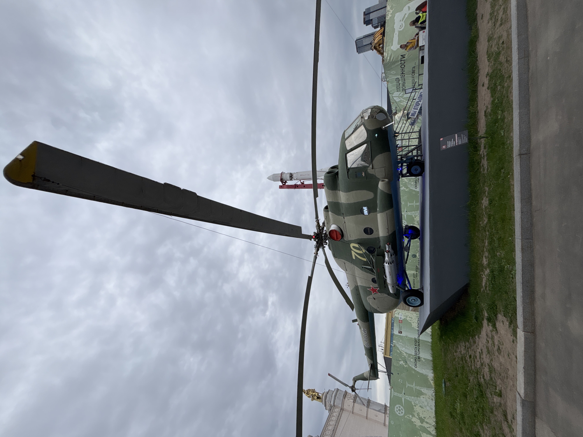 The image captures a Mil Mi-24 Hind attack helicopter on display, likely in Moscow, Russia. The helicopter is painted in a camouflage pattern, with the number "70" visible on its side and a red star insignia. It's positioned on a raised platform with wheels underneath. A missile is mounted on the wing pylon.
In the background, there are other structures, possibly exhibition halls, and a construction banner. There is an inscription on the lower right, containing Russian text. Above the helicopter, the sky is overcast. Part of a building with a golden sculpture can be seen in the lower center, indicating a potential landmark. The ground adjacent to the display is grassy with a paved walkway.