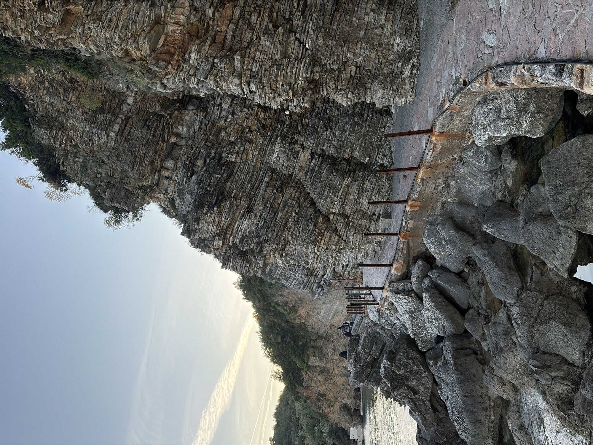 The image captures a dramatic coastal scene, likely in Budva, Montenegro, featuring a towering, stratified rock cliff face on the left, a narrow pathway running along its base, and the tranquil sea to the right. The cliff exhibits distinct horizontal layers of rock, with patches of sparse green vegetation clinging to its crevices and ledges.
A man-made pathway or walkway, constructed from stone or concrete, traces the contour of the cliff, providing passage along the waterline. Several rustic metal posts or supports extend from the pathway towards the water, possibly serving as a rudimentary railing or part of the structure. A black cable or pipe is also visible running along the cliff just above the path.
In the mid-ground, a small group of people are visible on the pathway and adjacent rocks, appearing to be seated or standing and observing the surroundings. Their activity suggests leisure or contemplation.
The time of day appears to be either early morning or late afternoon, indicated by the soft, warm light illuminating the sky. The sky transitions from a brighter, hazy hue near the horizon to a clearer blue above. A prominent, elongated contrail stretches across the sky, catching the sunlight. The sea is calm, with subtle reflections of the sky and light on its surface. The overall weather seems clear and serene. No visible text is discernible in the image.