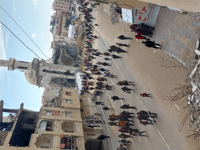 The image captures a wide street in Khan Yunis, Palestinian Territory, presenting a scene of extensive urban destruction amidst civilian activity. In the background, a prominent mosque with a light-colored dome and a tall, slender minaret exhibits severe damage, with large sections of its walls collapsed and interior structures exposed. To the left, several multi-story buildings, appearing to be residential or commercial, show significant structural damage, particularly on upper floors, featuring collapsed sections, exposed rebar, and charred areas. Balconies with debris or hanging items are visible.
The paved street is densely populated with hundreds of civilians, including adults and children, walking in various directions. They appear to be moving through the area, with no discernible organized activity. Their attire is civilian. In the middle distance, several white tents are erected amidst rubble near the damaged mosque, suggesting temporary shelters or aid provisions. Overhead utility lines crisscross the scene. In the immediate foreground, a pile of concrete debris and twisted rebar indicates the vantage point is from a damaged structure. On the right side of the street, a white banner with Arabic script, partially obscured by people, displays text including "شحن" (shipping) and "على مدار الساعة" (around the clock), suggesting a commercial service operating in the area. The sky is partially cloudy, indicating daylight.