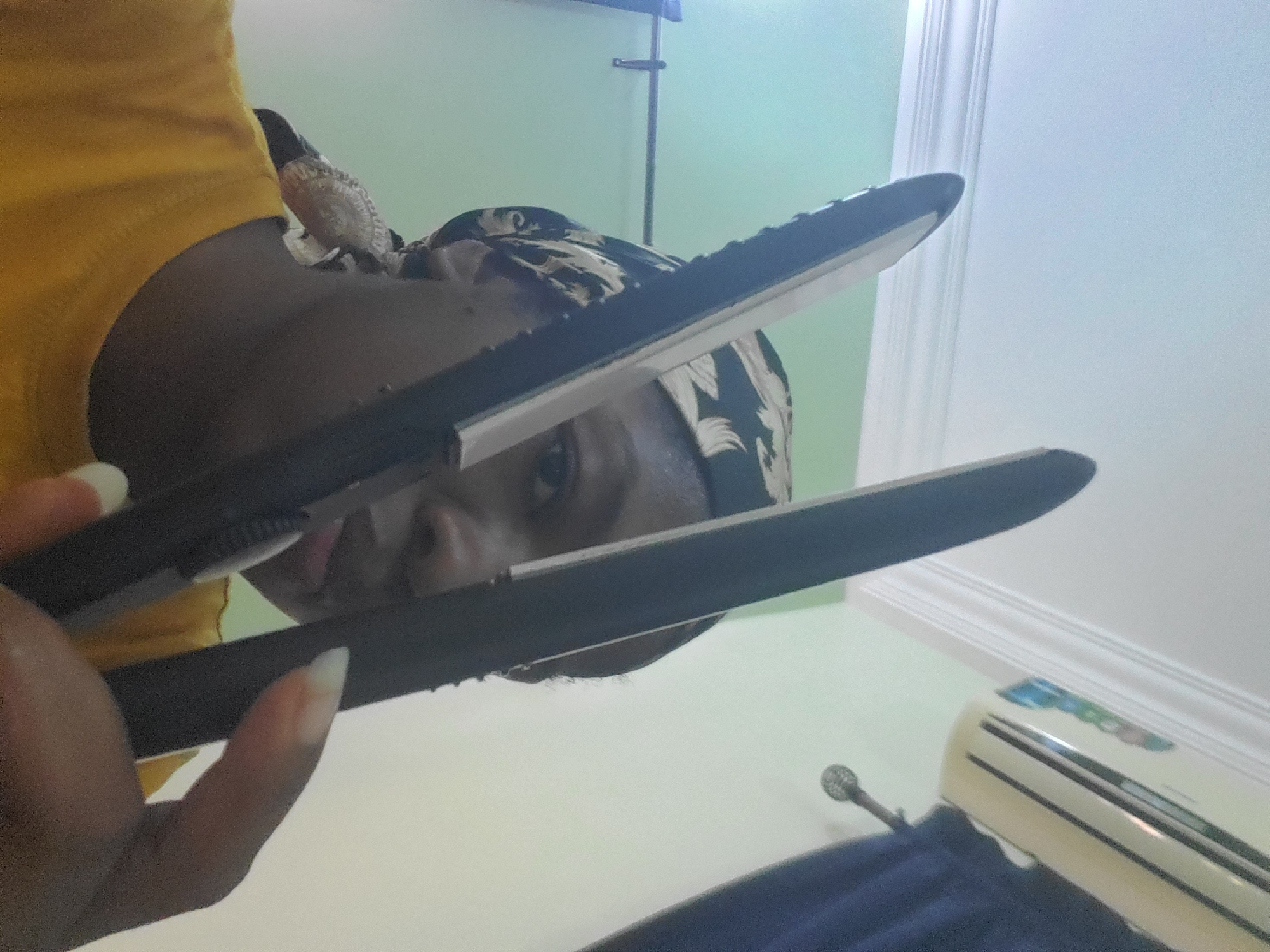 This image depicts an indoor scene with a person holding a hair straightener, positioned to partially obscure their face. The individual has dark skin and is wearing a yellow garment and a patterned head covering, which is black and white/cream. Their left hand, equipped with long, light-colored fingernails, grips the black hair straightener, which features metallic heating plates.
The background consists of light green walls and a white ceiling with decorative molding. To the upper right, a white or beige air conditioning unit is mounted on the wall, partially above a dark blue curtain. A metallic fixture, possibly a curtain tie-back or hook, is visible below the air conditioner. To the upper left, a metallic rod with a small clip or hook is present against the light green wall. The lighting appears uniform, suggesting an interior setting, possibly a room in a residence. The specific location in Barden Yana, Nigeria, as provided, cannot be independently verified from the visual information within the image.