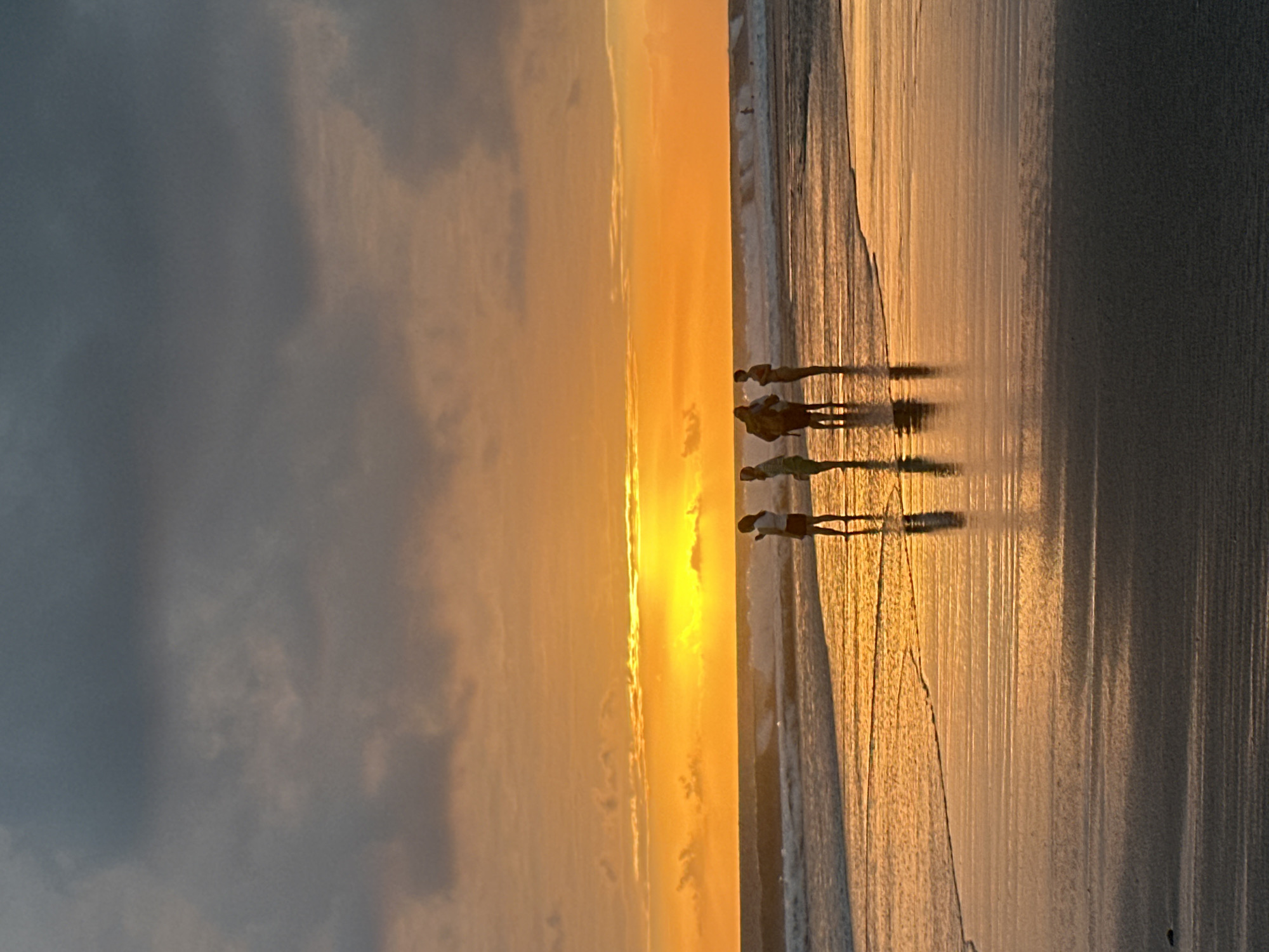 The image depicts a group of five silhouetted figures standing on a wet beach at sunset. The sky is filled with dramatic orange and yellow hues from the setting sun, with dark, brooding clouds overhead. The ocean is calm with gentle waves lapping at the shore, and the wet sand reflects the vibrant colors of the sky. The figures appear to be casually gathered, with some facing the ocean and others looking towards the horizon. The scene is serene and evokes a sense of peace and tranquility. The location is the coast of North Kuta, Indonesia.