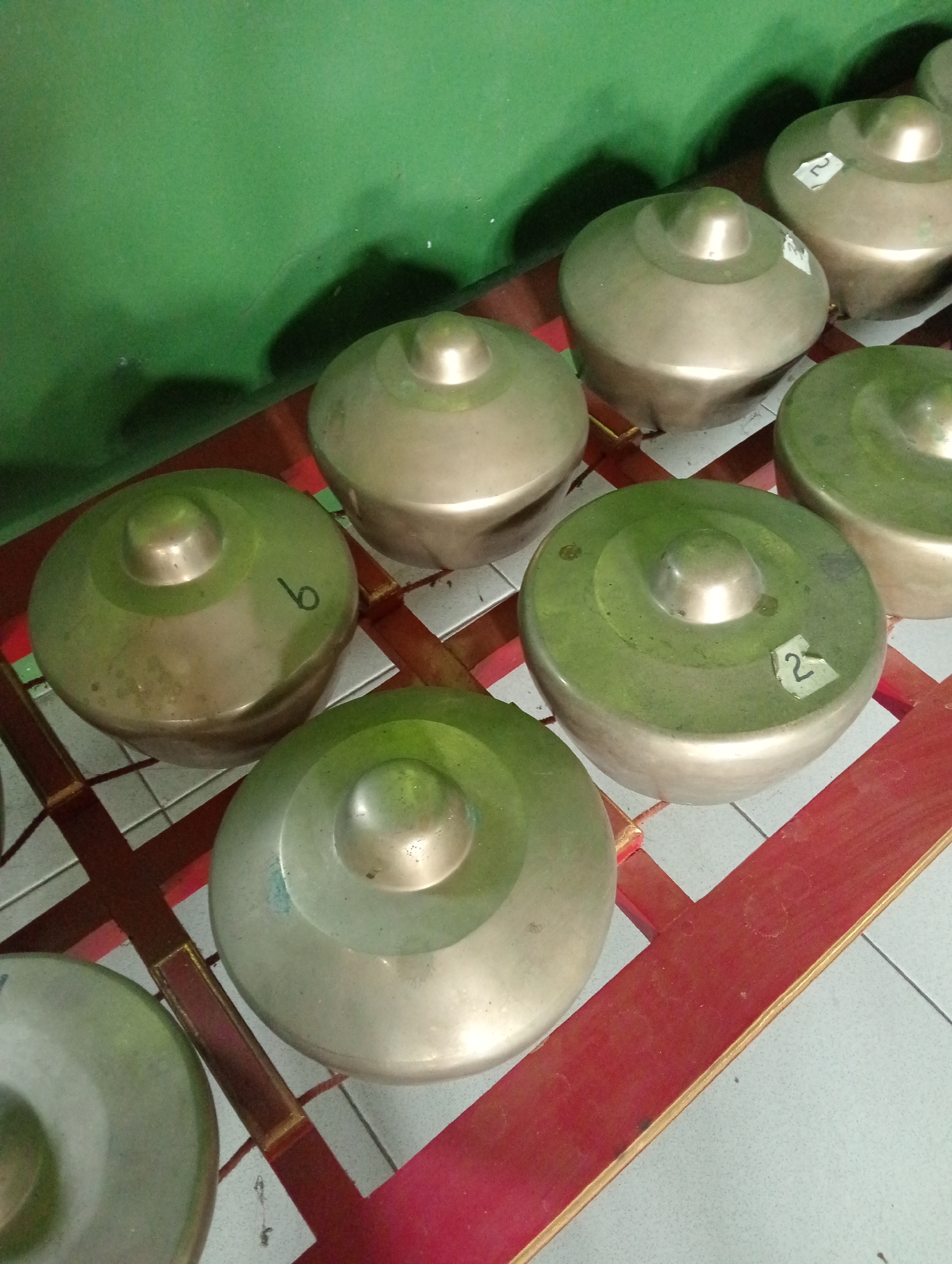 The image shows a collection of bronze gongs, likely part of a gamelan ensemble, arranged on a red wooden rack. The gongs are rounded with a raised central dome. Some gongs have numbers written on them in black ink, such as '6' and '2'. The gongs are positioned on a light-colored tiled floor, with a green wall in the background. The lighting suggests an indoor setting. The scene is likely in Demak, Indonesia, given the context of gamelan music being a traditional Indonesian art form often found in cultural centers and musical institutions. There are no people visible in the image. The overall impression is one of musical instruments prepared for use or storage.