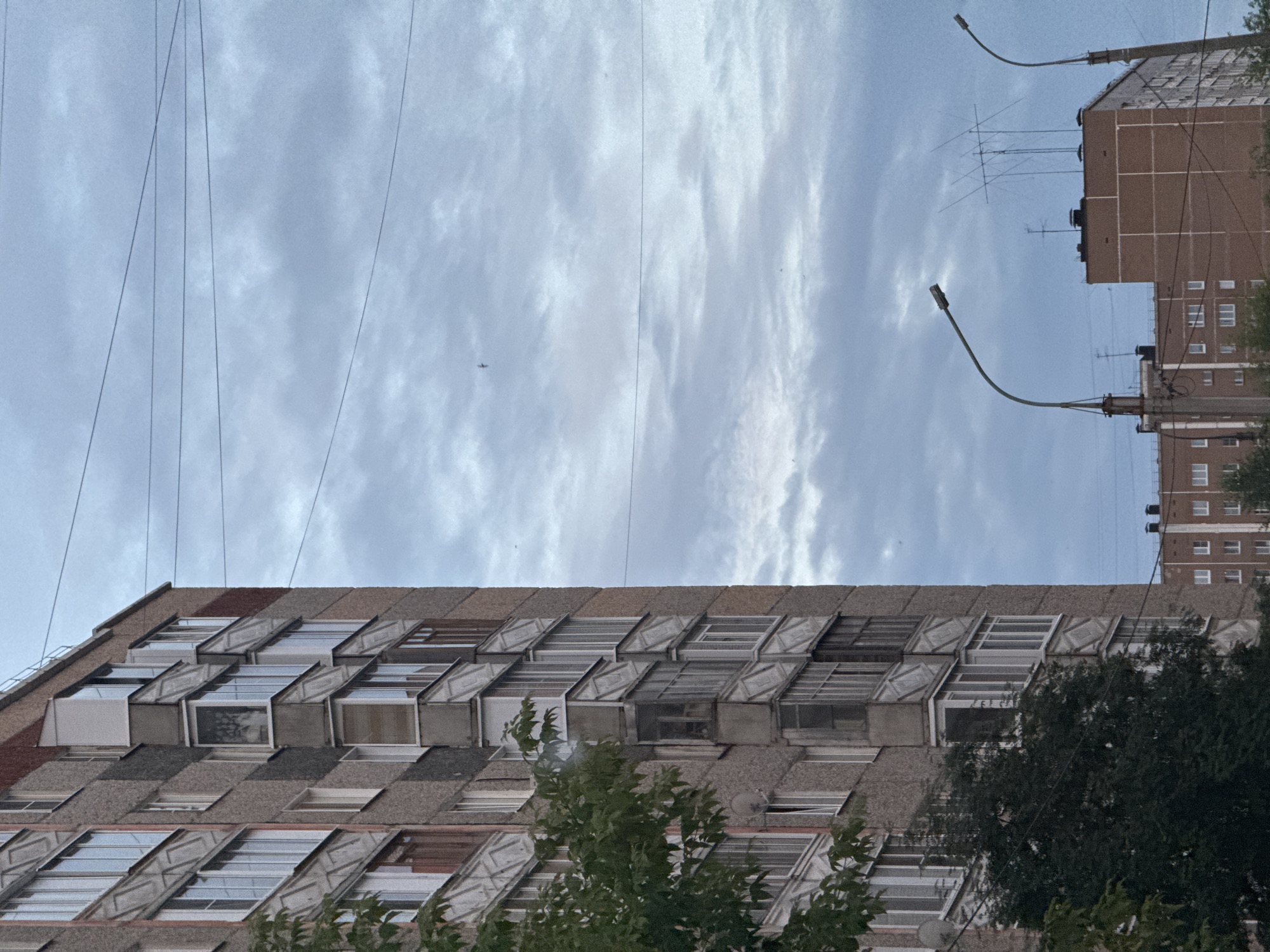 The image depicts a view of an apartment building in Izhevsk, Russia, under a cloudy sky. The building on the left is a multi-story residential structure with a grey, textured facade and numerous balconies. The building on the right is a brick structure with white window frames. Electrical wires are visible crisscrossing the sky, and two streetlights are present in the lower portion of the frame. There are no people or animals visible in the image. The scene appears to be a typical urban environment.