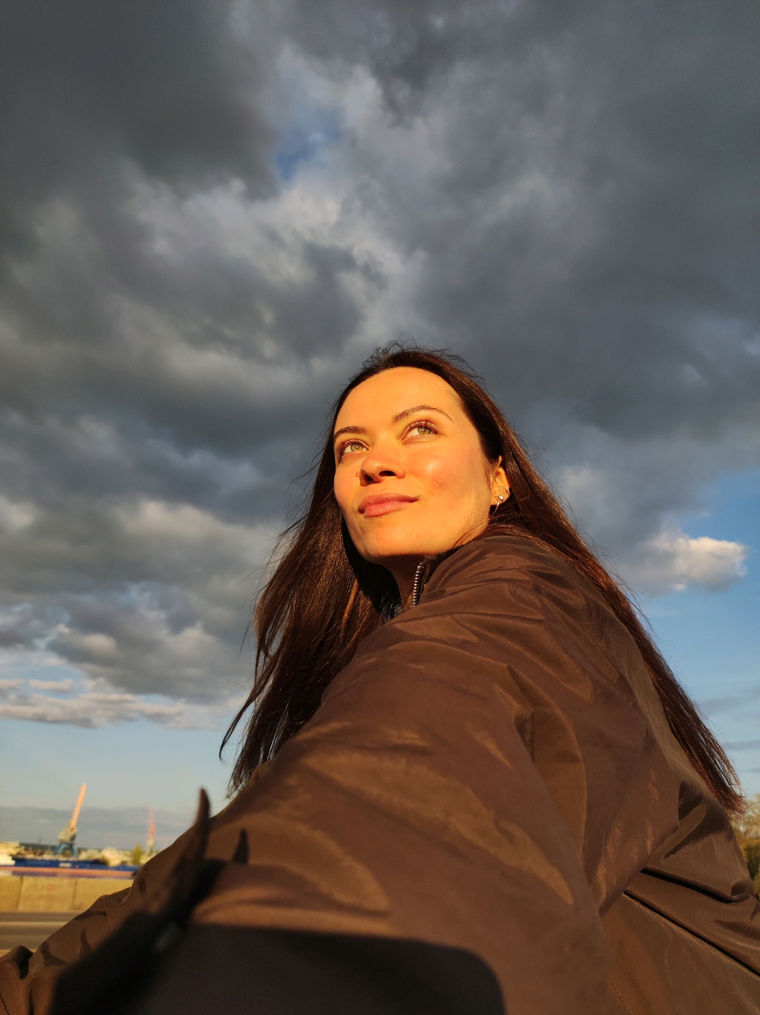 A low-angle medium shot captures a woman looking upwards and to the right against a dramatic sky filled with dark, swirling clouds and patches of blue sky. The golden hour light illuminates her face, highlighting her features and the sheen on her skin. She has long, dark hair that flows behind her. She is wearing a dark, possibly brown, jacket that covers her from the chest down. In the background, a hazy industrial landscape with cranes is visible under the cloudy sky, suggesting a port or industrial area. The lower part of the image shows a blurred road or pathway.