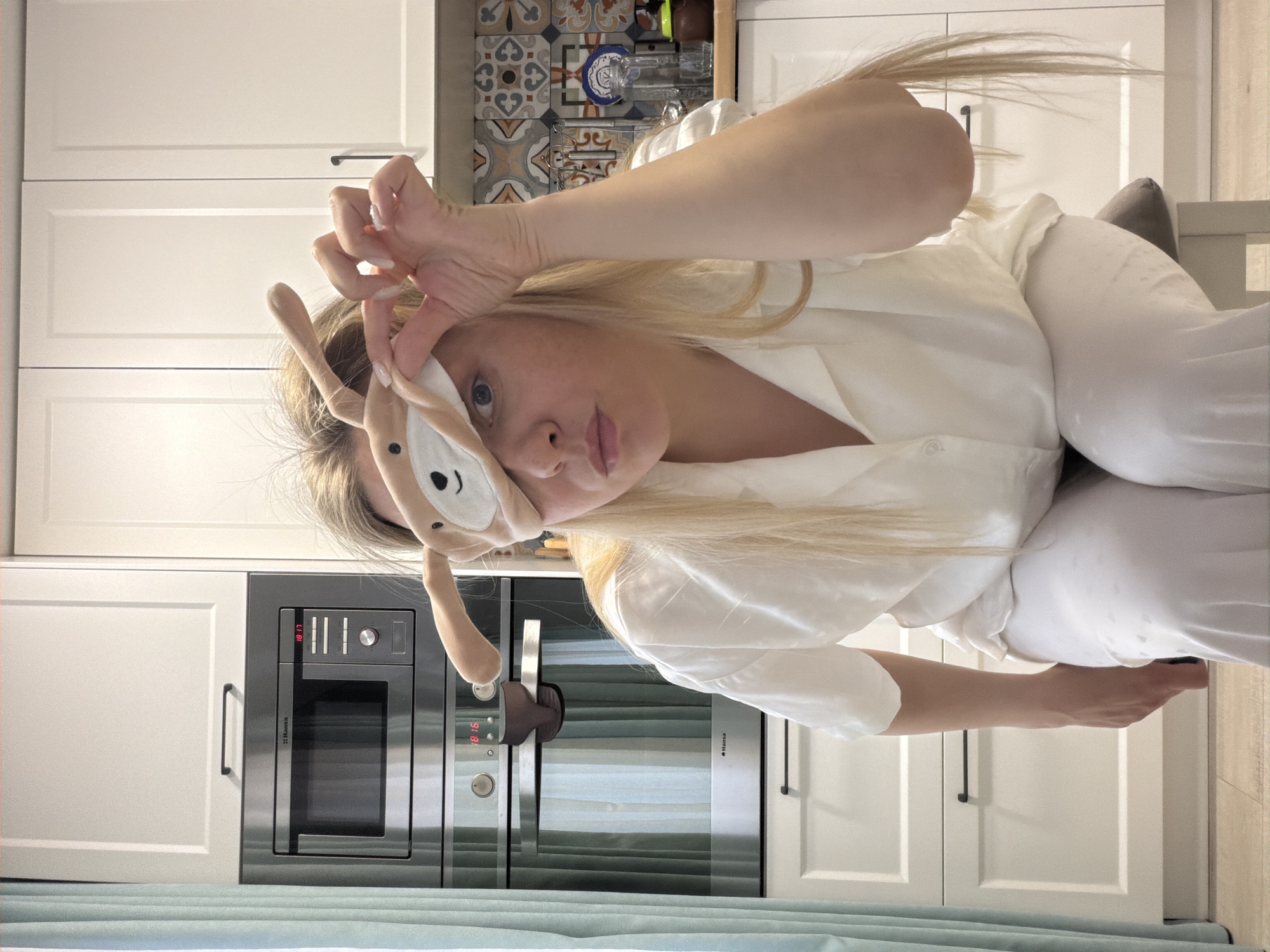 A young woman is captured in a playful, casual moment within what appears to be a modern kitchen setting in Votkinsk, Russia. She is shown from the waist up, with long blonde hair draped over her shoulders. She wears a loose-fitting, light-colored, possibly white or cream, button-up shirt and matching pants.
Her face is partially obscured by a beige, animal-themed sleep mask, which features soft, floppy ears and an embroidered face, resembling a dog or bunny. She holds the mask up with her left hand, revealing her blue eyes, while her right hand forms a "framing" gesture near her face. Her lips are slightly parted, and she has light-colored nail polish on her manicured nails. The woman's expression is gentle and engaging.
The background shows a contemporary kitchen with white, flat-panel cabinets adorned with minimalist black handles. Above the lower cabinets, a backsplash features decorative ceramic tiles with a geometric pattern in tones of blue, brown, beige, and white. A built-in stainless steel oven or microwave, branded "Hansa", is prominent, displaying the time "18:17" in red digital numbers. Part of a light blue or teal curtain or fabric is visible in the lower foreground. To the right, a wooden floor and a grey upholstered chair or stool can be seen. The overall impression is bright, clean, and personal.