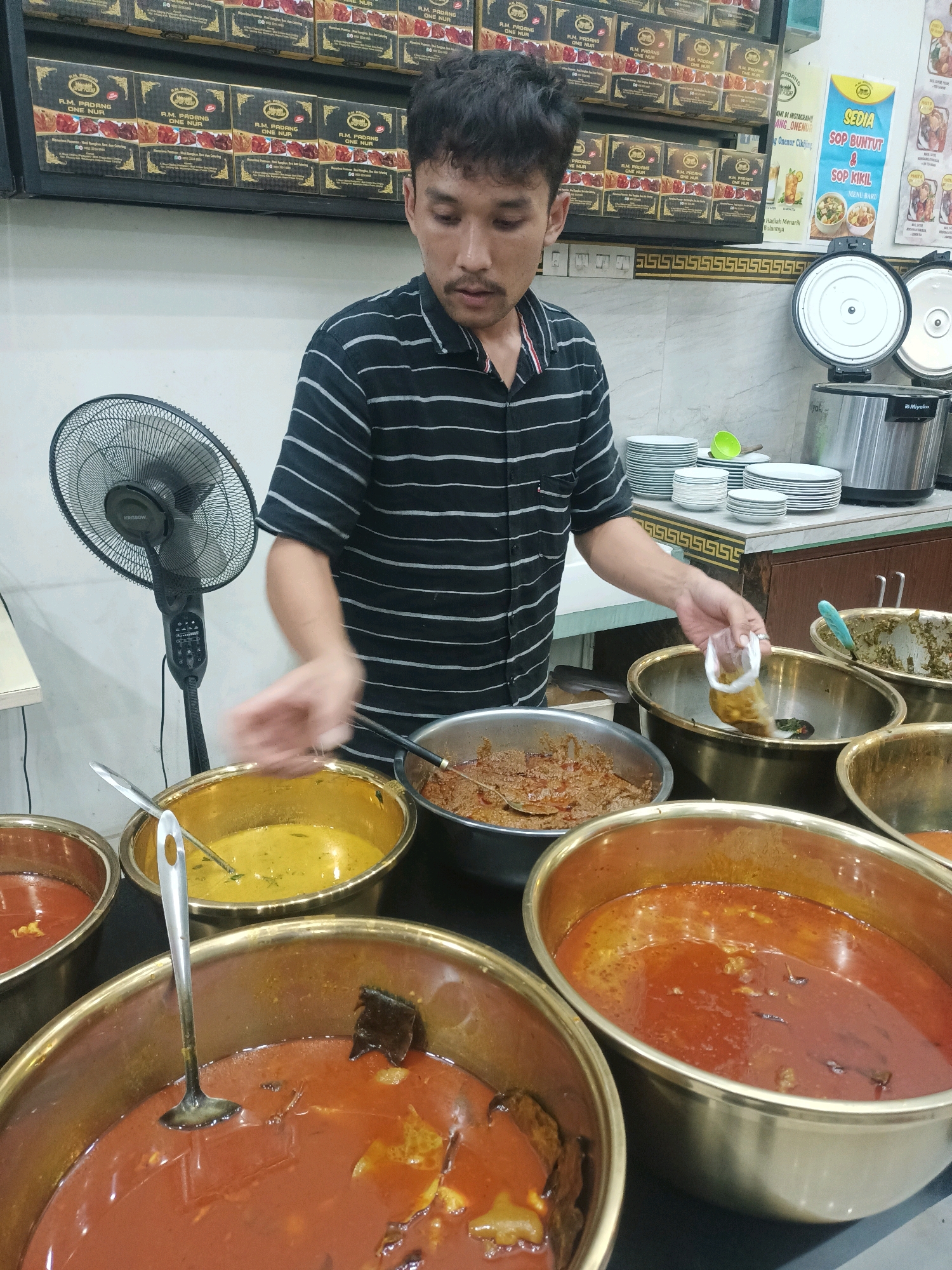 In a food stall located in Sukamukti, Indonesia, a man wearing a black and white striped shirt is standing behind a counter filled with various dishes. He is holding a small, clear plastic bag containing what appears to be some sort of condiment. In front of him are several large, golden-colored bowls containing different types of curry or stew. To his left, a black oscillating fan is visible. Behind the man, shelves are stacked with boxes of what seem to be food items, and a banner advertising "Sedia Sop Buntut & Sop Kikil" is displayed. A rice cooker and stacks of plates are also present on the counter.