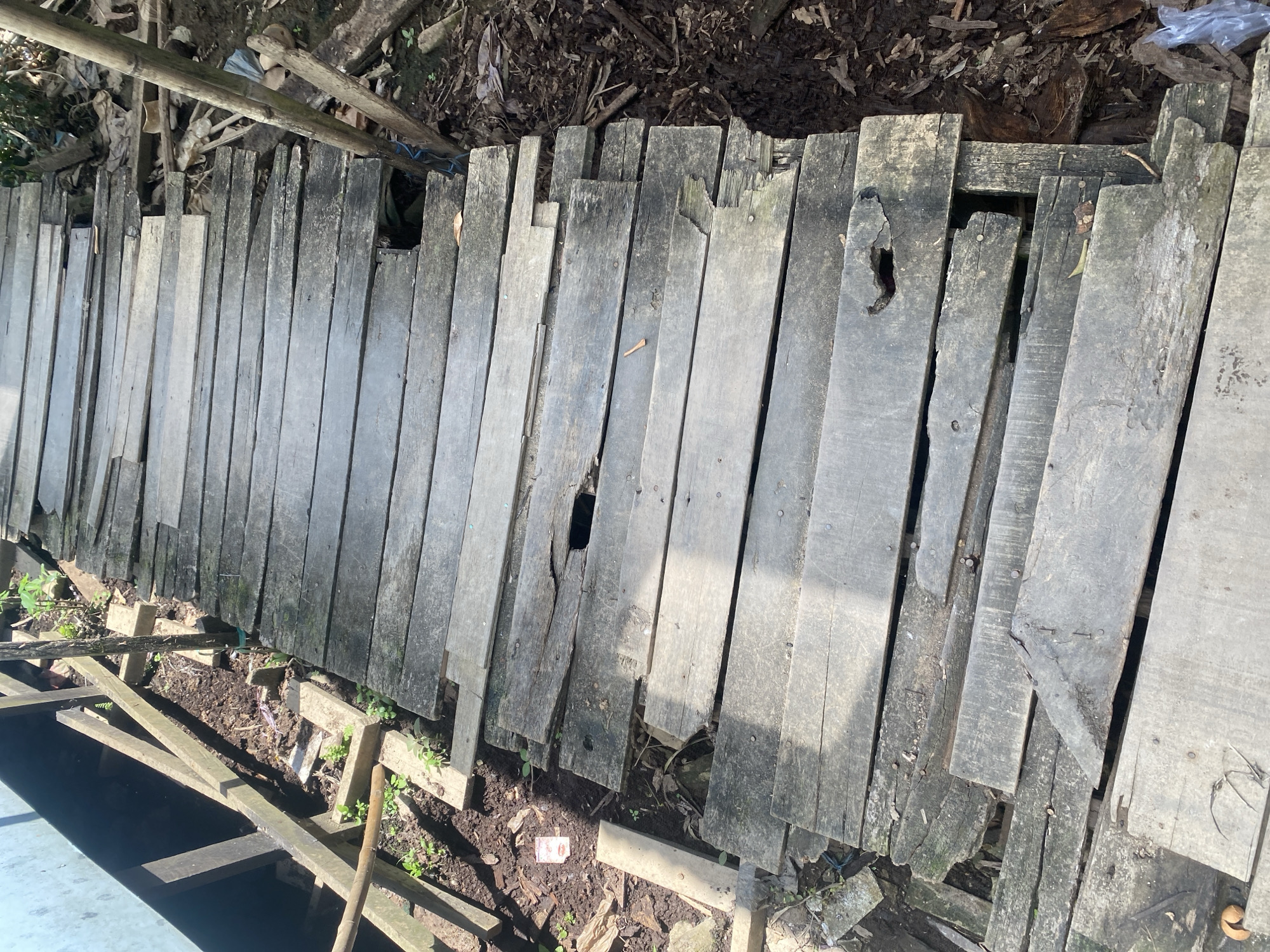 The image depicts a weathered wooden walkway in Berangas Timur, Indonesia. The walkway, constructed from aged, gray planks, shows signs of significant decay and damage. Several planks are broken, missing, or warped, creating gaps and uneven surfaces. The walkway is bordered by a dirt path with scattered debris, including leaves, plastic, and plant growth. Supporting wooden beams are visible beneath the walkway, some of which also appear damaged. The overall scene suggests an unmaintained or deteriorating pathway in an outdoor, possibly rural or semi-rural, environment.
