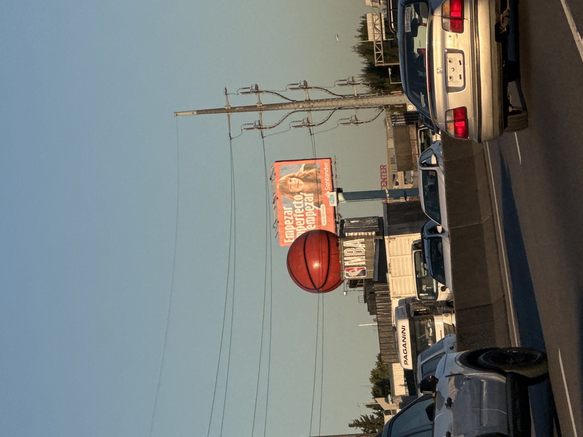 The image, taken sideways, captures a roadside scene near Martínez, Argentina, dominated by a large inflatable basketball with an NBA logo on top of shipping containers, placed next to an advertising billboard for Santander bank. The billboard shows the slogan "Empezar perfecto, empezar", and a woman's face, and the logo of "PC" is also visible. The setting is a busy highway with multiple vehicles in the traffic. The sky above is clear and light blue. Power lines extend over the road. Some of the vehicles are identified as a Honda Accord and a Paganini truck.