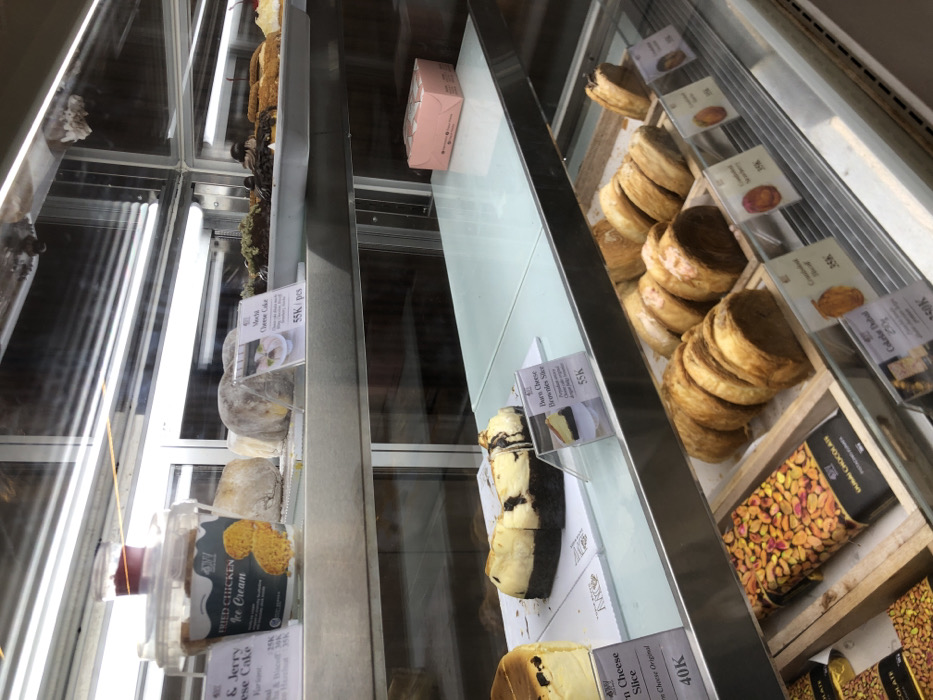 This image captures a close-up view of a multi-tiered refrigerated glass display case, likely inside a bakery or dessert shop in Panjang, Indonesia.
The top shelf, from left to right, features a transparent plastic bucket labeled "Fried Chicken Ice Cream" with "Jerry & Jerry Cheese Cake" branding and various prices (28K-35K IDR), topped by a small, clear container with red liquid. Next to this are two round, light-colored, flour-dusted mochi-style cakes. An acrylic standee labels "Mochi Cheese Cake" at 55K IDR per piece, displaying an image of a pink dessert. Further right are assorted pastries, some with green toppings, others with chocolate glaze.
The middle shelf showcases a partially sliced cheesecake with a dark brown base and light-colored top, resting on a white rectangular board. Adjacent is an acrylic standee for a "Burn Cheese Brownies Slice," priced at 55K IDR. The rest of this shelf is empty, save for a small pink rectangular box positioned in the back right corner.
The bottom shelf displays multiple chocolate bars with pistachio toppings, including one marked "DARK CHOCOLATE PISTACHIO," arranged in wooden trays. To their right are rows of golden-brown, possibly croissant-like, laminated pastries, also in wooden trays. Price labels for "Croissant Original," "Croissant Chocolate," and "Croissant Strawberry" are visible at 35K IDR each, alongside a label for "Cokelat Dubai 250gr" at 150K IDR.
The display case itself is made of glass and stainless steel, with bright fluorescent lighting illuminating the products from above. No individuals or active interactions are present in the frame.