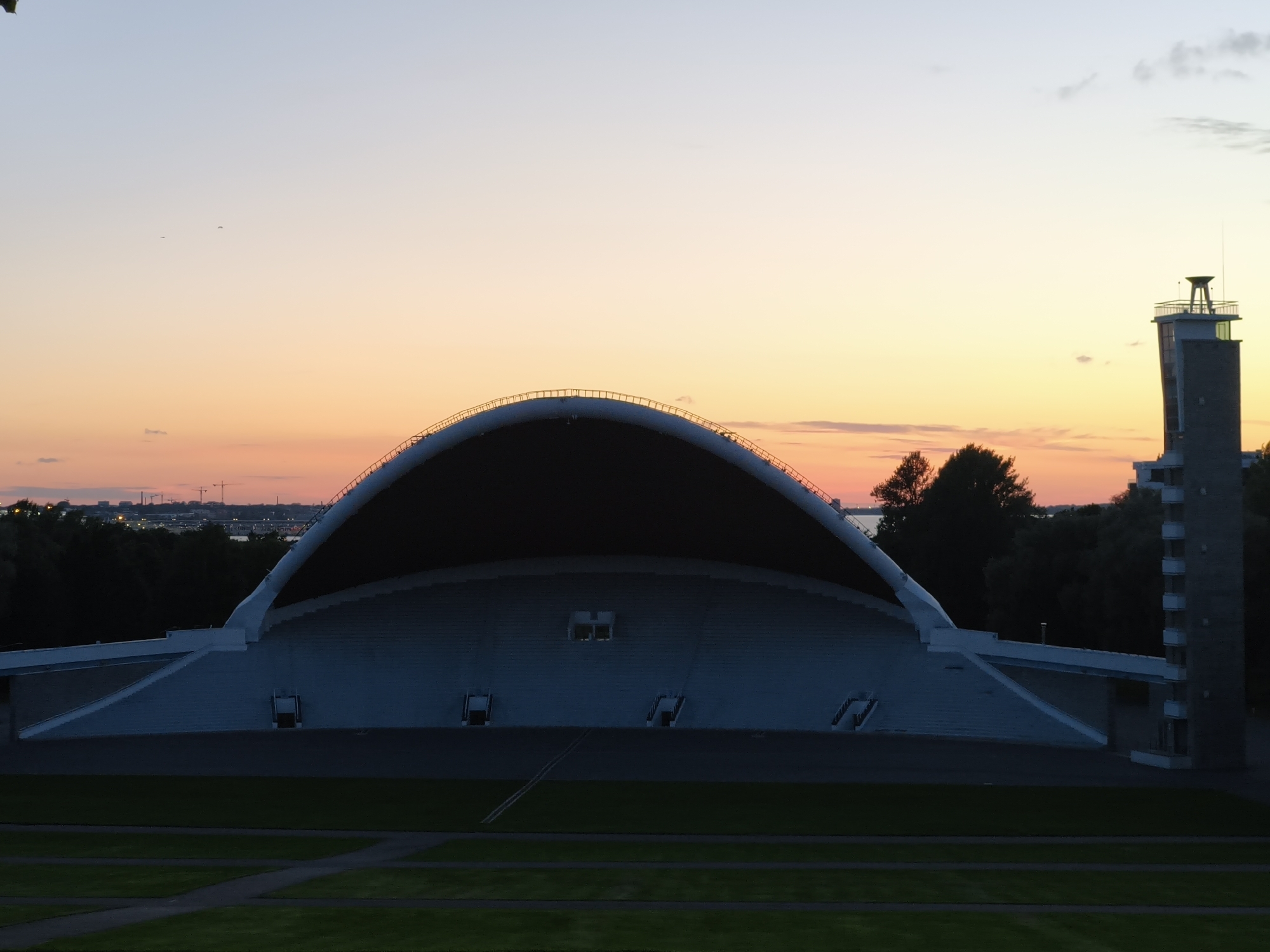 An aerial view of the Tallinn Song Festival Grounds in Tallinn, Estonia, captures the iconic amphitheater against a pastel sunset. The grand, shell-shaped structure, made of white concrete, dominates the scene, its tiered seating receding into shadow. To the right of the amphitheater stands a tall, grey tower with a white, open-air platform at its summit. Green grass fields with paved pathways are visible in the foreground, with dark trees forming a line behind the amphitheater. The sky transitions from a pale blue at the top to soft orange and pink hues near the horizon, with faint silhouettes of cranes and buildings suggesting a distant cityscape. A few birds are visible as tiny dots in the sky. The overall mood is serene and tranquil, bathed in the soft light of dusk.