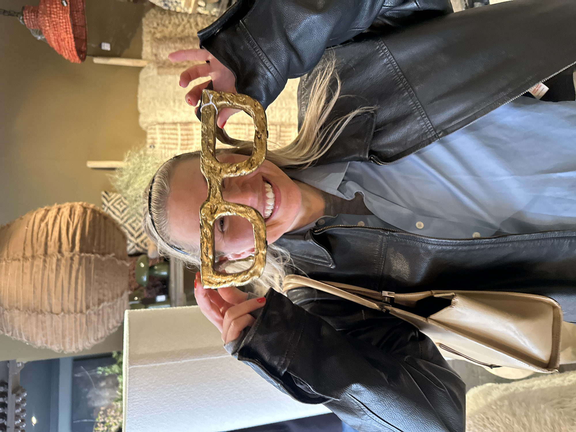 A woman with blonde hair and red nails, wearing a black leather jacket and a light blue shirt, is holding up oversized, gold-colored letter D-shaped spectacles in front of her face. She is smiling. A tan purse with a silver closure is slung over her shoulder. The background is a retail environment, featuring light fixtures, various textured items, and shelving. Due to the lack of definitive landmarks, the location cannot be definitively identified as London, United Kingdom.