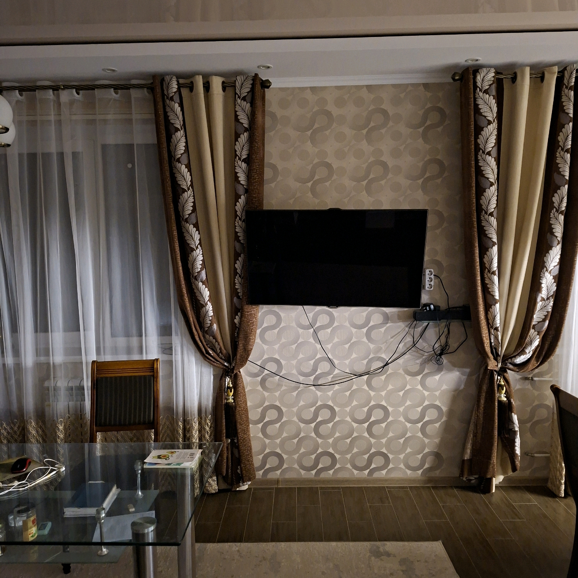 The image shows an interior scene, possibly a living room, in Anapskaya, Russia. The room features patterned wallpaper, floor-to-ceiling curtains on either side of a television mounted on the wall, and a glass-top table with various objects on it. A wooden chair is partially visible behind the table. The overall impression is one of a lived-in space, with no people present in the frame.