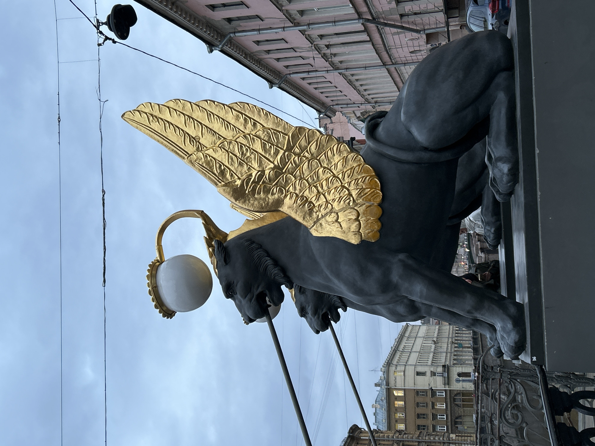 The image displays a majestic outdoor scene featuring the iconic winged lion statues of the Bank Bridge in Saint Petersburg, Russia. Two large, dark grey griffin-like statues are prominent, with one in the foreground capturing most of the attention. Each statue boasts elaborate golden wings, intricately detailed with feathered patterns. From the head area of each statue, a golden, curved arm extends, holding a spherical white lamp, serving as an ornate lamppost for the bridge.
The perspective is upward-looking, showcasing the grandeur of the statues against an overcast, light grey sky. Above the statues, a series of overhead wires and a black, bell-shaped fixture are visible, part of the urban infrastructure. In the background, classic European architecture lines the street, with multi-story buildings featuring light-colored facades, numerous windows, and decorative cornices. Some windows in the distant buildings show faint illumination, suggesting a dim, possibly late afternoon or early evening sky, though the overall brightness points to a cloudy day. A decorative metal railing, part of the bridge, is partially visible beneath the statues. The scene is static, with no human figures or specific activities depicted. There is no visible text in the image.