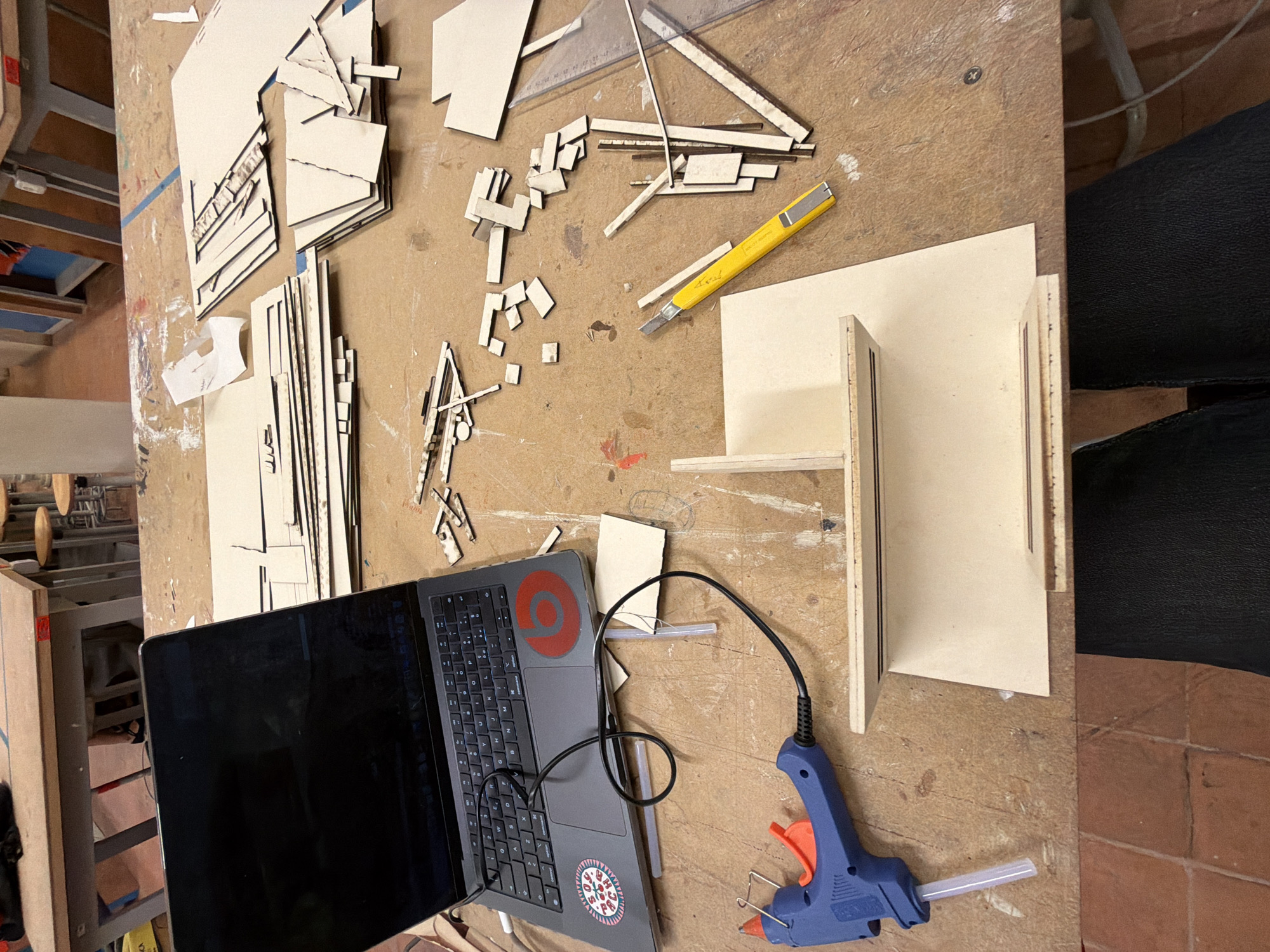 A cluttered workbench is covered with unfinished architectural model components, including laser-cut pieces of cardboard, a partially assembled model, a laptop, a glue gun, and a utility knife. The laptop has stickers on it, including a red circle and a round multicolored sticker. The workbench is stained and scratched. The floor is tiled with red terracotta tiles. Part of a black upholstered seat is visible on the right side. The objects suggest a workshop or design studio environment. There is no evidence of a specific location within Milan, Italy.