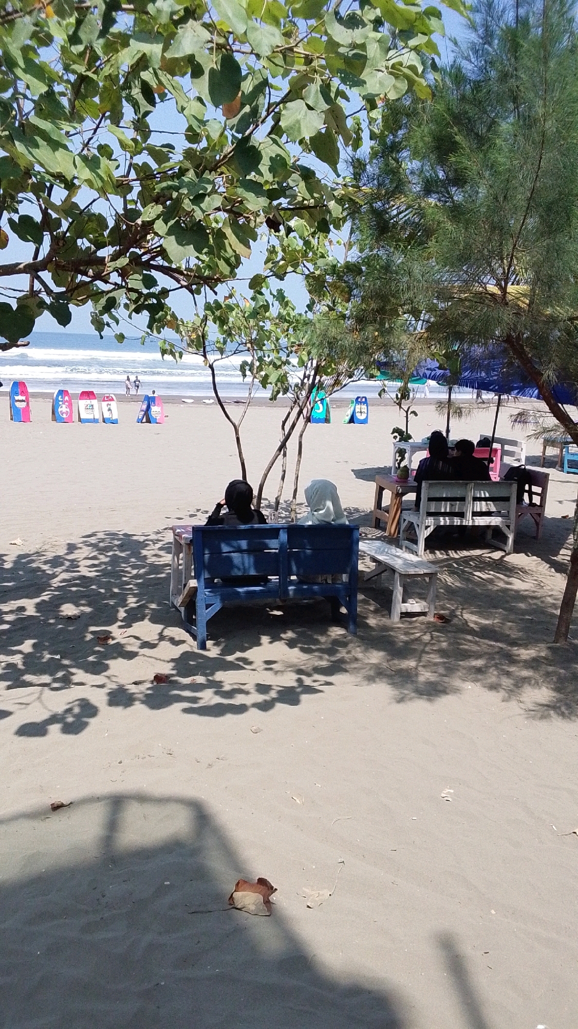 This image depicts a beach scene at Desa Pagandaran, Indonesia, on a sunny day. In the foreground, two people are seated on a blue wooden bench, facing away from the camera towards the ocean. The person on the left is wearing a black headscarf, and the person on the right is wearing a white headscarf. They are positioned under the shade of a tree with green leaves, with shadows of the branches cast on the sand. To the right of the blue bench, there is a small white wooden table.
Further into the background, other people are seated at tables under blue umbrellas, suggesting a seating area for beachgoers. Several colorful surfboards are standing upright in the sand in the middle ground, arranged in a line. The ocean is visible in the distance, with waves gently breaking on the shore. The sand is light brown, and there are a few fallen leaves scattered on the ground. The overall environment is that of a relaxed beach atmosphere.