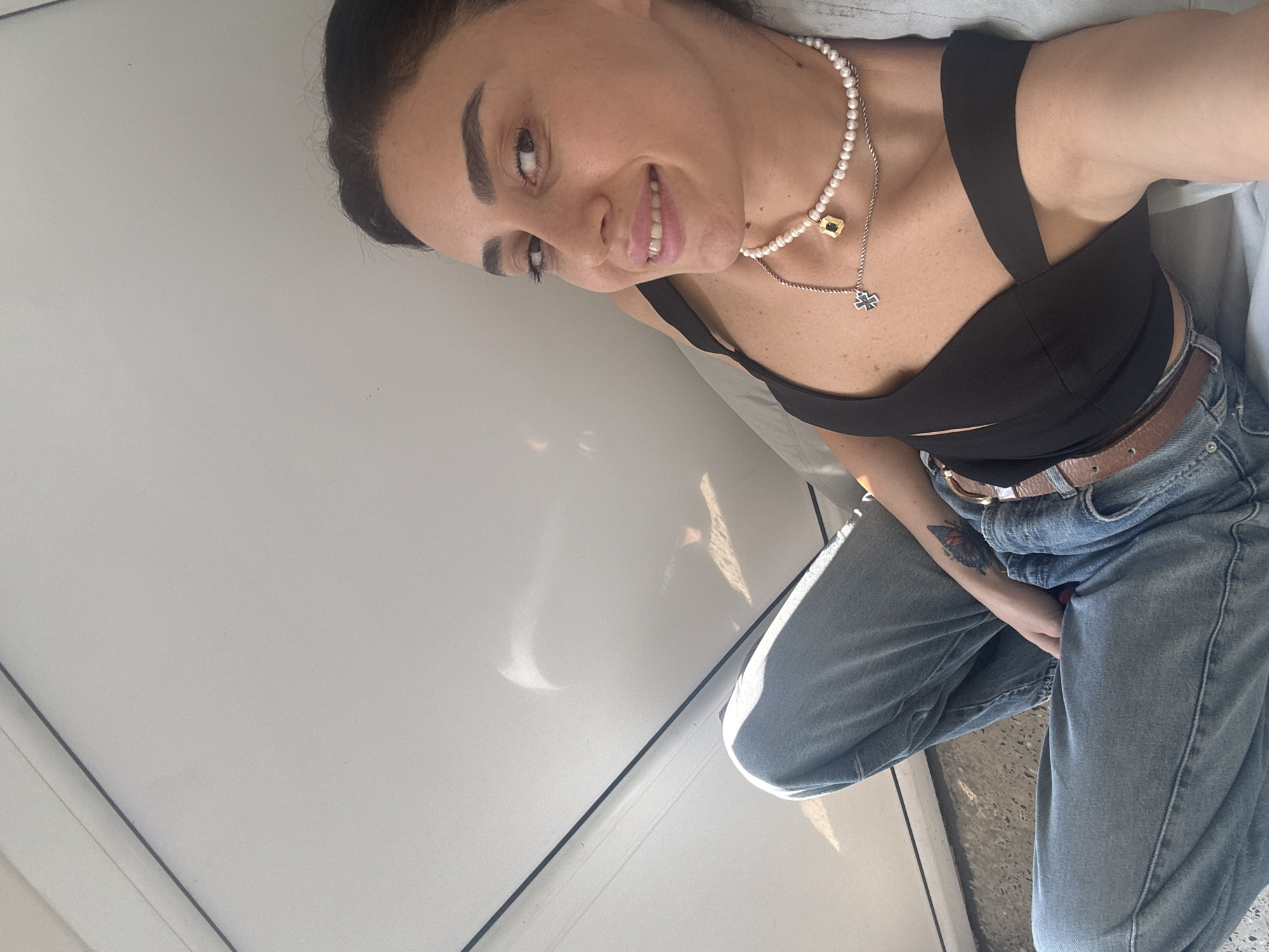 A woman is reclining against a white, slightly reflective surface, possibly the exterior of a building or a large piece of furniture. She is wearing a black crop top with a crisscross design over the bust, light-wash denim jeans, and a brown belt. She has dark hair pulled back, and is wearing a pearl necklace with a pendant and a second, thinner chain with a cross-shaped pendant. A tattoo of a butterfly is visible on her left forearm. The lighting suggests an outdoor setting, with strong sunlight casting shadows on the white surface and on her body. The ground appears to be concrete or gravel. The location is identified as Tbilisi, Georgia.
