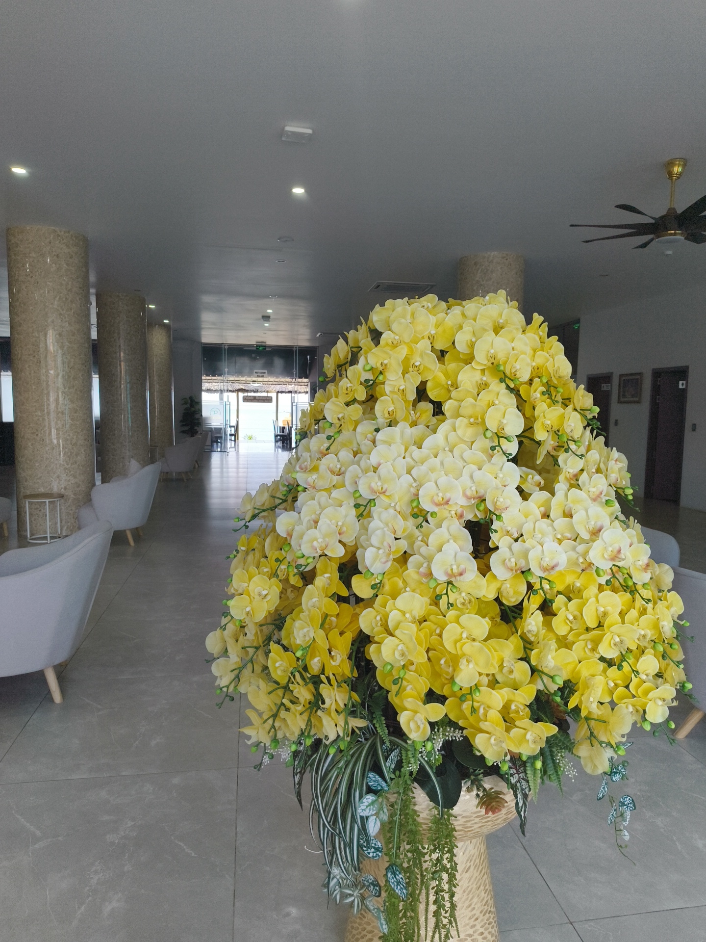 The image shows an interior shot of a hotel lobby in Phu Thuy Ward, Vietnam. In the foreground, a large artificial arrangement of yellow and white orchids is prominently displayed in a woven basket. The lobby features marble-patterned floor tiles and large, round marble-clad columns. To the left, several light gray armchairs are arranged, and a small side table with a white top is visible. In the background, through a glass-enclosed entryway, an outdoor area with tables and chairs can be seen, suggesting a restaurant or seating area. Further down the hallway, two doors are visible on the right wall, along with framed pictures. A ceiling fan is mounted on the ceiling in the upper right portion of the image. The lighting is bright and consistent throughout the space.