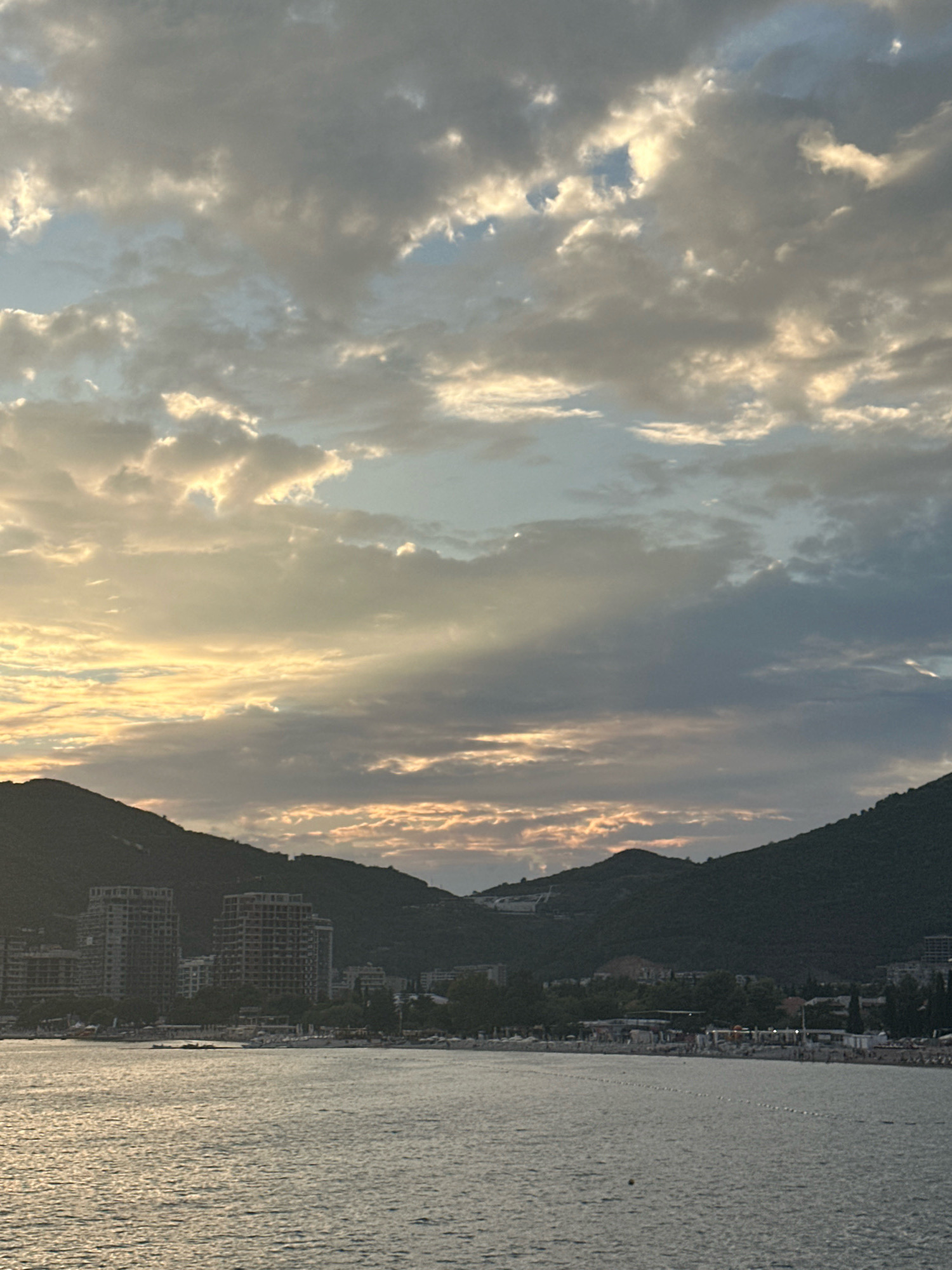 The image depicts a coastal cityscape at dusk in Budva, Montenegro. The foreground features a calm body of water, likely the Adriatic Sea, reflecting the muted light of the setting sun. Along the shore, a beach lined with umbrellas and lounge chairs is visible, with several people scattered across it, suggesting a leisurely atmosphere. Behind the beach, a line of trees separates the coast from buildings.
Dominating the midground and background are the imposing silhouettes of dark, tree-covered hills that frame the city. Nestled between these hills, and partially obscured by the foliage, are clusters of buildings, including several multi-story structures that appear to be hotels or apartment complexes, some of which seem to be under construction or renovation, as indicated by exposed frameworks.
The sky above is a dramatic canvas of clouds, with patches of soft sunlight breaking through, creating ethereal rays that illuminate the upper atmosphere. The overall mood is serene and picturesque, capturing the transition from day to night in a coastal Mediterranean setting. The scene captures the interplay between natural landscape and urban development, characteristic of popular tourist destinations like Budva.