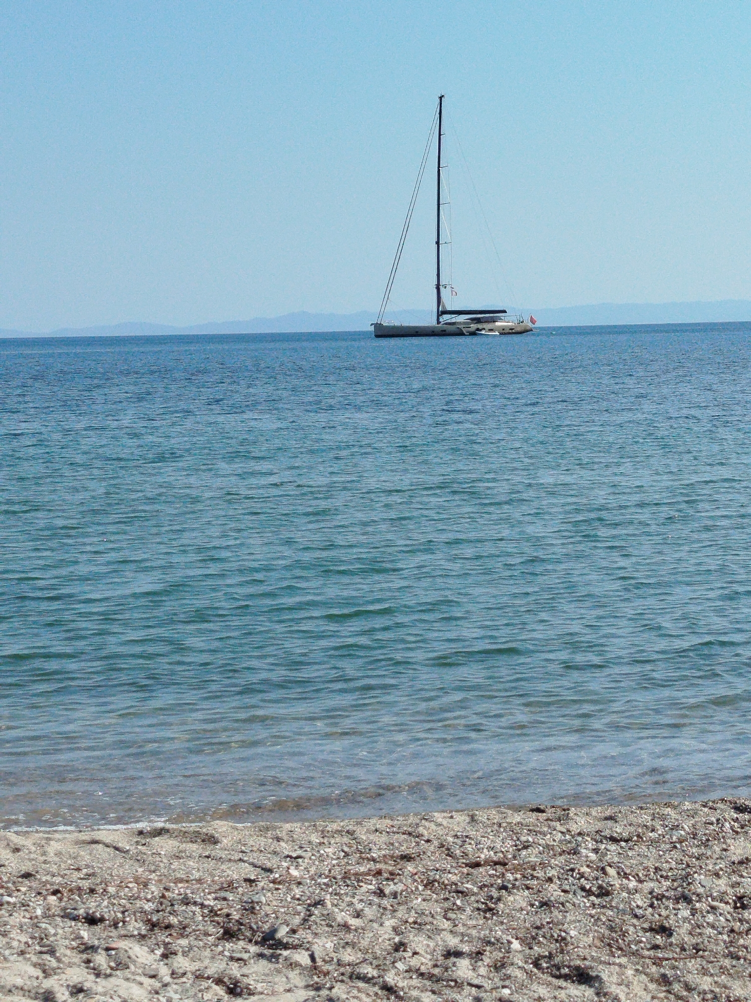 A sailboat is anchored in the blue sea off the coast of Sithonia, Greece. The sailboat has a tall mast and appears to be a catamaran. The water is calm with small waves. In the background, there are faint mountains visible on the horizon. The foreground shows a sandy beach with small stones and debris. The sky is clear and blue.