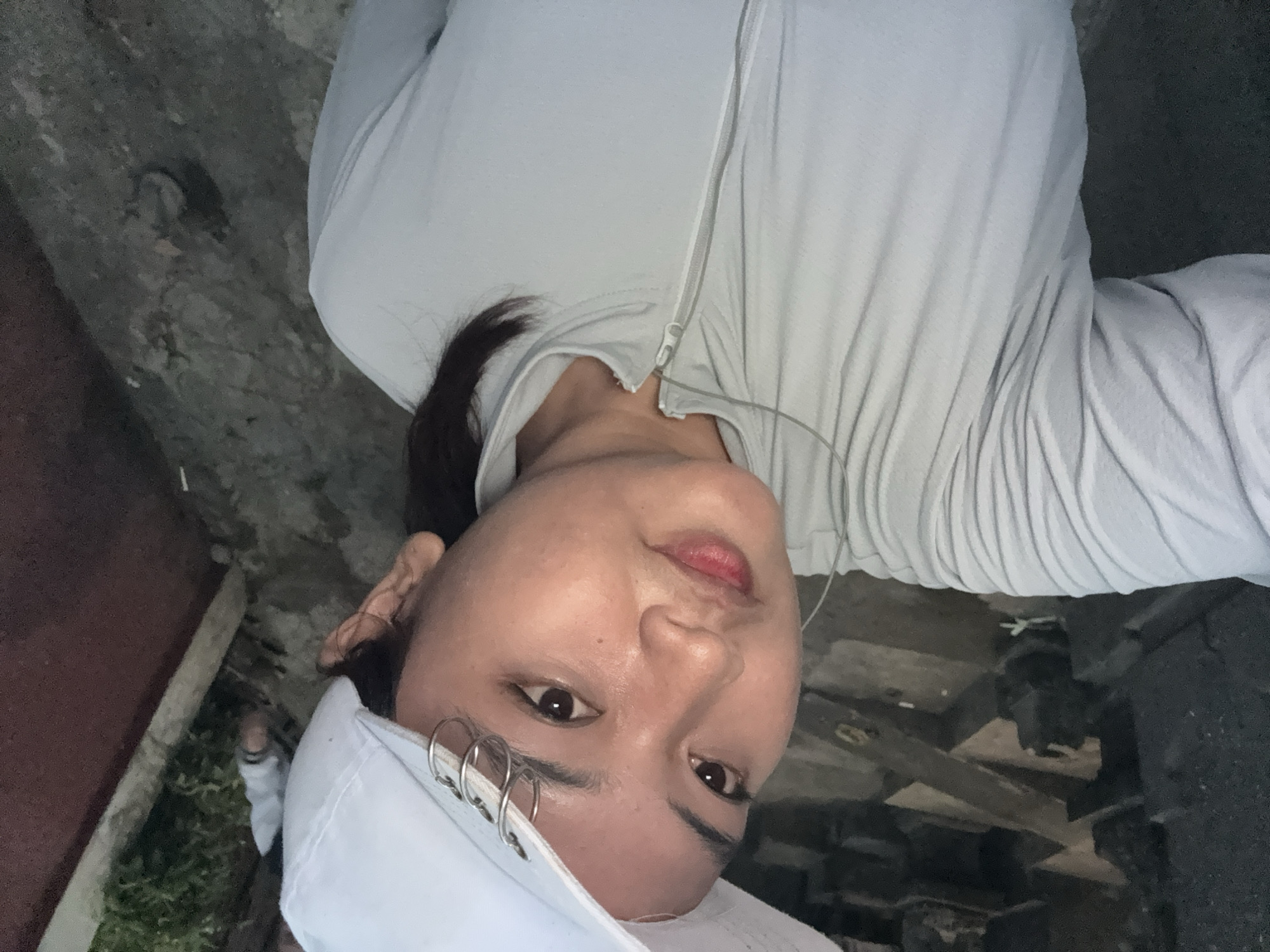In an outdoor setting with stone structures and foliage, a young woman with dark hair, wearing a white baseball cap and a light gray zip-up jacket, takes a selfie. The cap has several silver hoop earrings attached to its brim. She is looking at the camera with her head tilted. The background suggests an outdoor pathway or courtyard, possibly with stairs. The lighting is dim, indicating it may be dusk or early evening. A second person, partially visible in the background, is wearing a similar light-colored jacket.