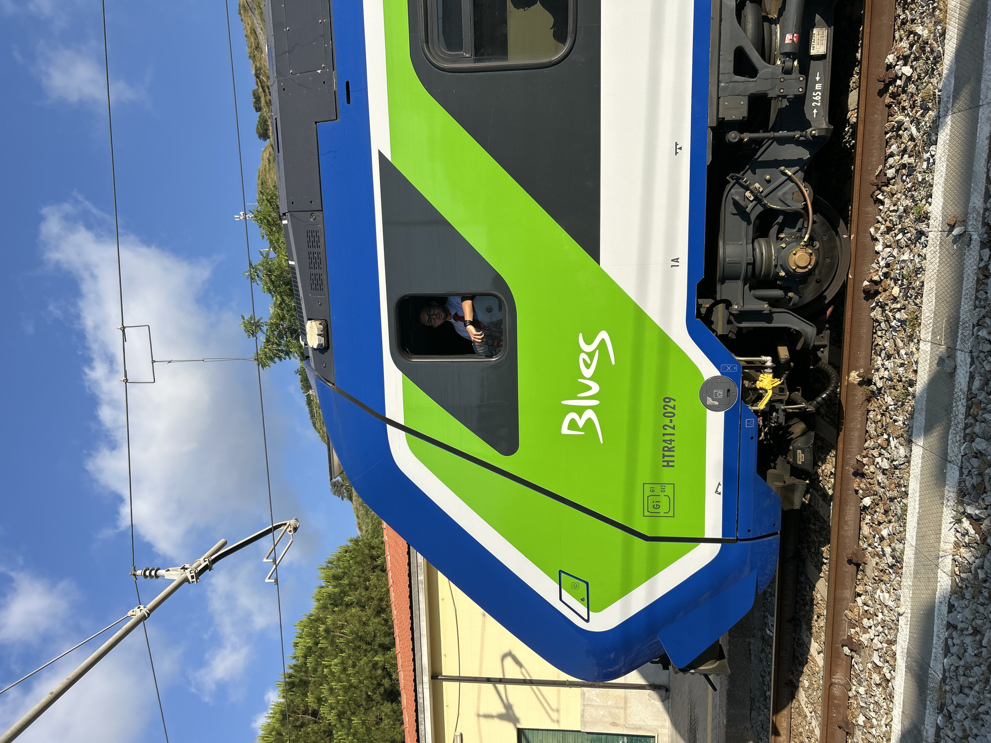 The image shows the side of a blue and green train, labeled "Blues," at a station in Tropea, Italy. A young man wearing glasses and a red tie is visible in the train's window, appearing to be the driver. The train is on tracks, with a yellow building and trees in the background. The sky is blue with some clouds. The train has the number HTR412-029 printed on its side. The image captures a moment of travel or transit, with the train poised for departure or arrival.