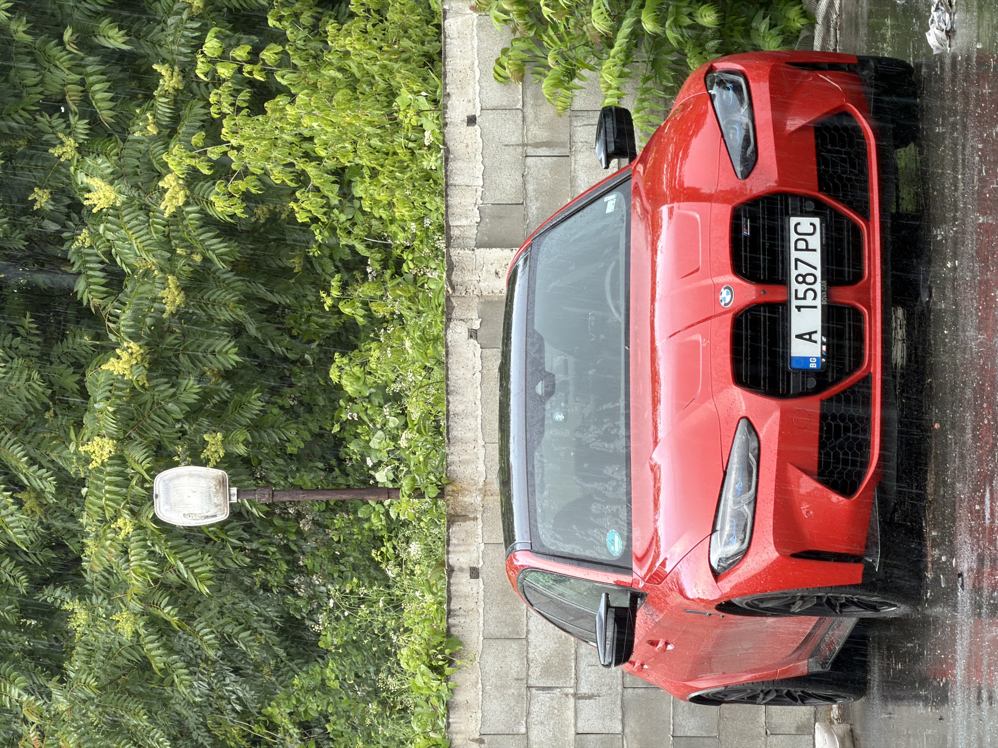 The image shows a red BMW M3 sedan parked in front of a concrete block wall. The car's front license plate reads "BG A 1587 PC". Behind the wall, lush green foliage is visible, and a lamppost with a transparent cover stands amongst the plants. It is raining, and raindrops are visible in the air and on the car and ground, creating reflections on the wet asphalt. The scene appears to be in an outdoor parking area, likely in Burgas, Bulgaria, given the license plate prefix "BG". No people are visible in the frame.