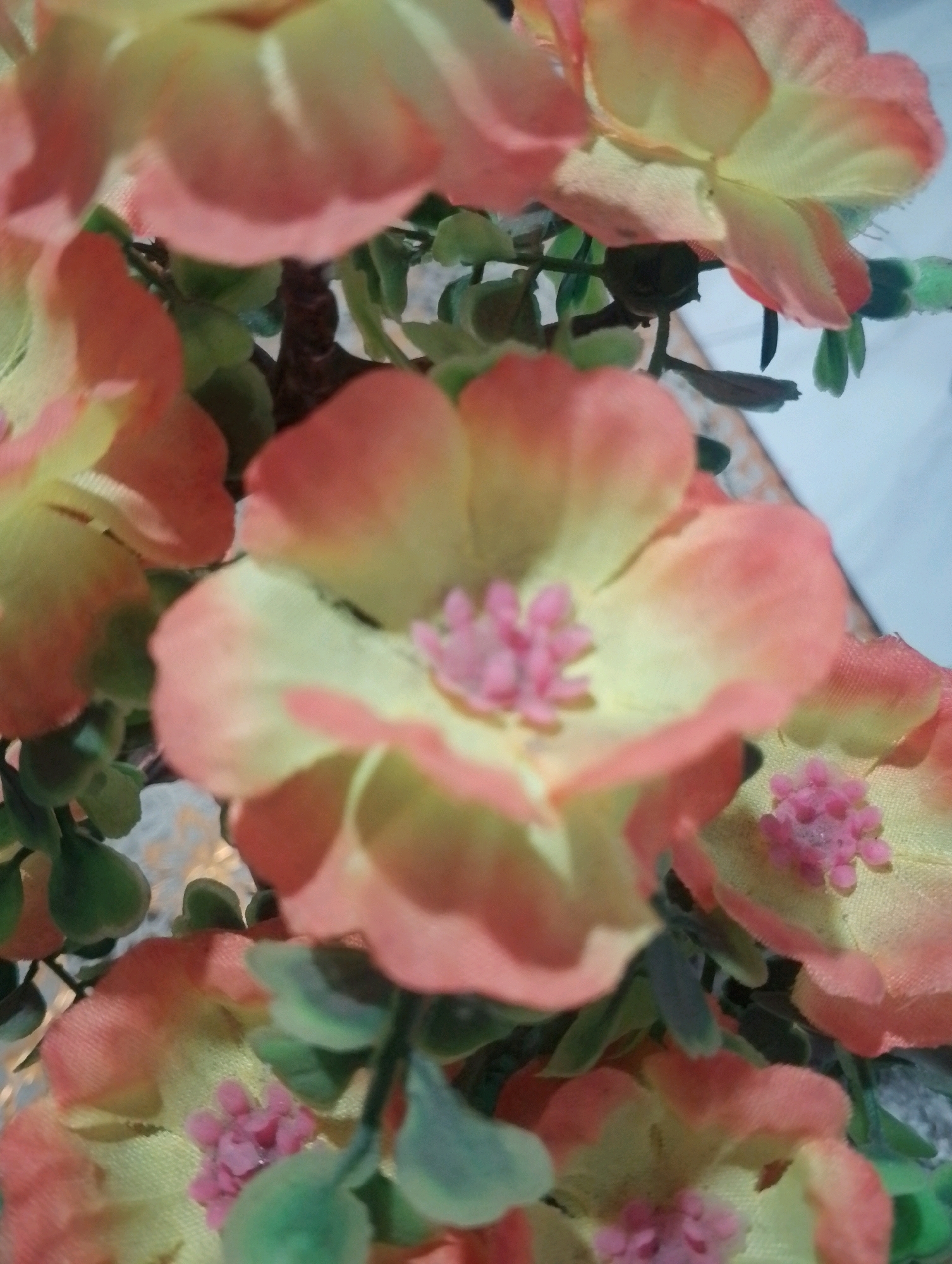 A close-up, blurred shot of artificial flowers in various shades of pink and yellow, with green leaves. The flowers appear to be silk or fabric and are arranged in a cluster, possibly as a decorative arrangement. The lighting is soft, creating a gentle gradient on the petals. The background is out of focus, showing a light-colored surface and hints of darker tones. There are no people or any identifiable environmental elements that would suggest a specific location in Doko, Indonesia, or anywhere else. The image focuses solely on the texture and color of the artificial flowers.