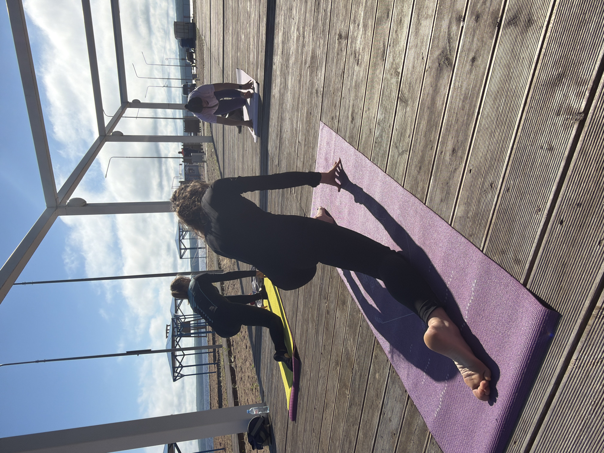 In Chaikovsky, Russia, on a sunny day, three women are practicing yoga on a wooden boardwalk next to a beach. The boardwalk is adjacent to a body of water, with a sandy beach area to the left. In the background, there are some small blue gazebos and utility poles with lights. The sky is bright blue with scattered white clouds.
The woman in the foreground is on a purple yoga mat, performing a low lunge pose with her right leg forward and her left leg extended behind her, with her toes on the mat. She is wearing black leggings and a black long-sleeved top.
To her left, another woman is on a yellow yoga mat, also in a low lunge position, with her left leg forward and her right leg extended behind her. She is also wearing black athletic wear.
Further back, a third woman is on a yoga mat in a tabletop position, with her hands and knees on the mat. She is wearing a light pink t-shirt and dark leggings.
A water bottle and a bag are placed on the sand near the woman on the yellow mat. The scene appears to be an outdoor yoga class or personal practice session.