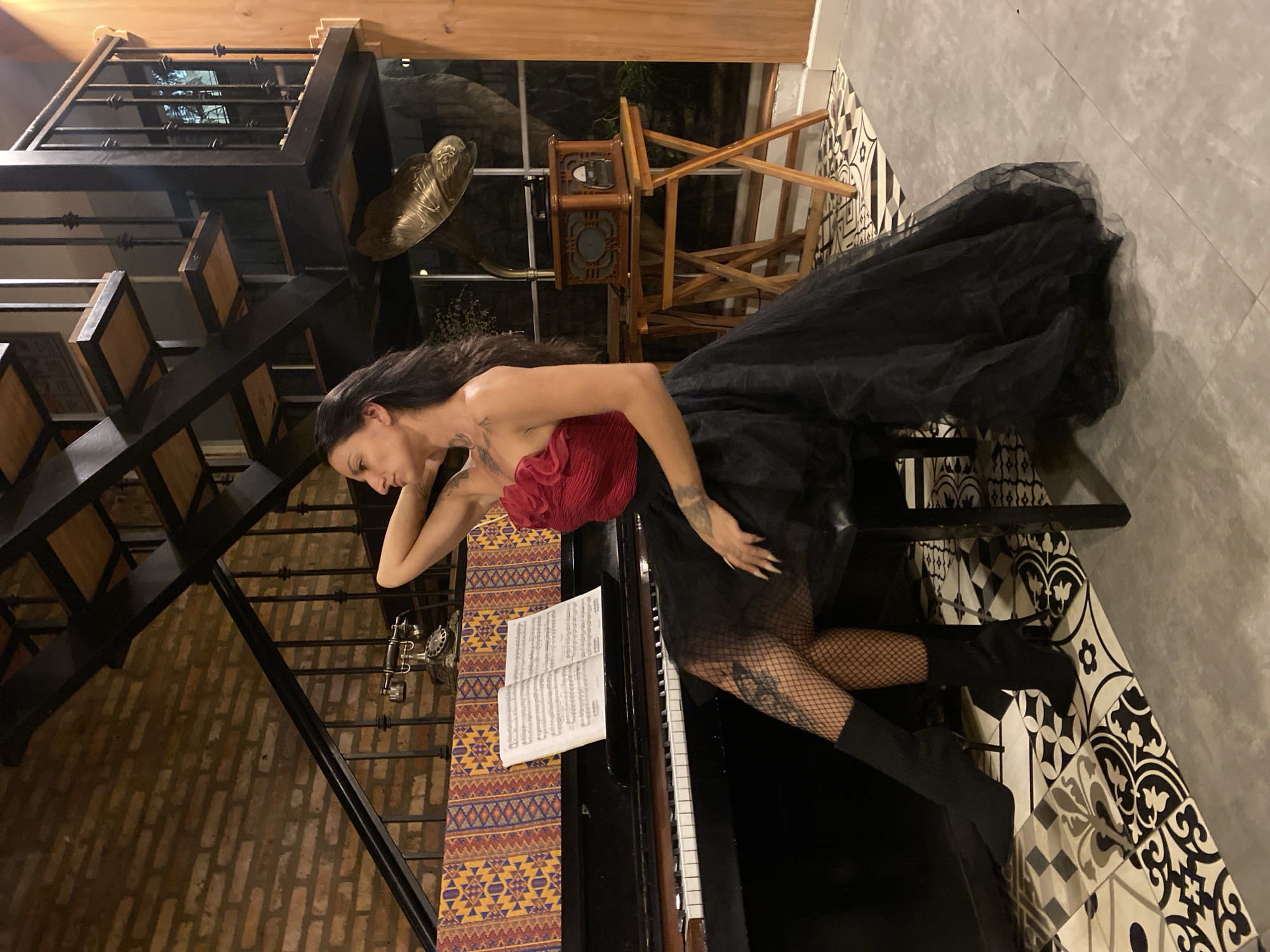 A woman with long, dark hair and tattoos on her arm and leg sits sideways on a stool in front of a black piano. She wears a strapless red top with ruffled detail and a voluminous black tulle skirt. Her legs are clad in black fishnet stockings and black pointed-toe boots. The piano has sheet music open on its stand, and a decorative fabric with geometric patterns is draped behind it. In the background, a black metal staircase with wooden steps ascends. To the right of the piano, a vintage gramophone with a wooden base sits on a foldable wooden table. The scene is set indoors, with a brick wall and wooden elements visible. The floor is tiled with a black and white patterned design. The location is identified as Da Lat, Vietnam.