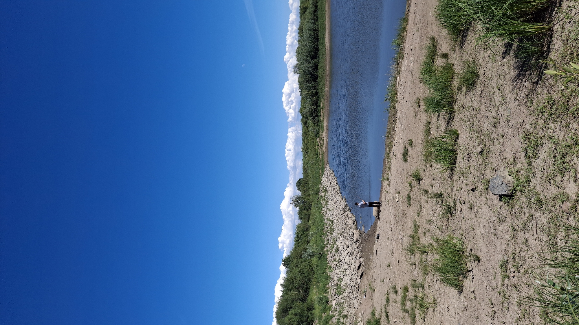 A person stands on the sandy shore of a lake, fishing. The water is a deep blue, reflecting the bright blue sky. The shore is lined with rocks and sparse green grass. In the background, a dense line of trees and bushes forms a natural barrier. The sky is clear with a few wispy white clouds on the horizon. The image appears to be taken on a sunny day. The location is the city of Kaporskoe, Russia.