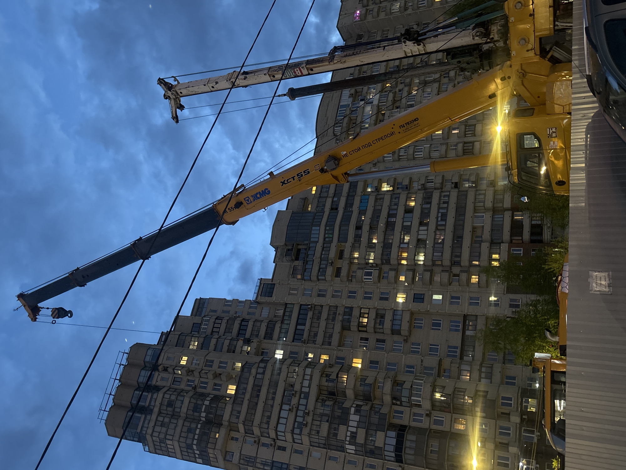 The image shows a low-angle view of two cranes in front of a tall apartment building at dusk or dawn. The building has numerous windows with lights on in some of them. The cranes, one yellow and the other white, are extended, with their booms angled towards the sky. Power lines are visible in the foreground. Text is visible on the yellow crane, indicating that it is an XCMG XCT55 S, with a warning in Russian. A car is partially visible on the right side of the image. The building's architecture and the Russian text suggest the location is likely in a Russian city. The distinctive architectural style of the building resembles Soviet-era apartment buildings found in many cities across Russia, but without specific markers, Saint Petersburg cannot be confirmed as the location.